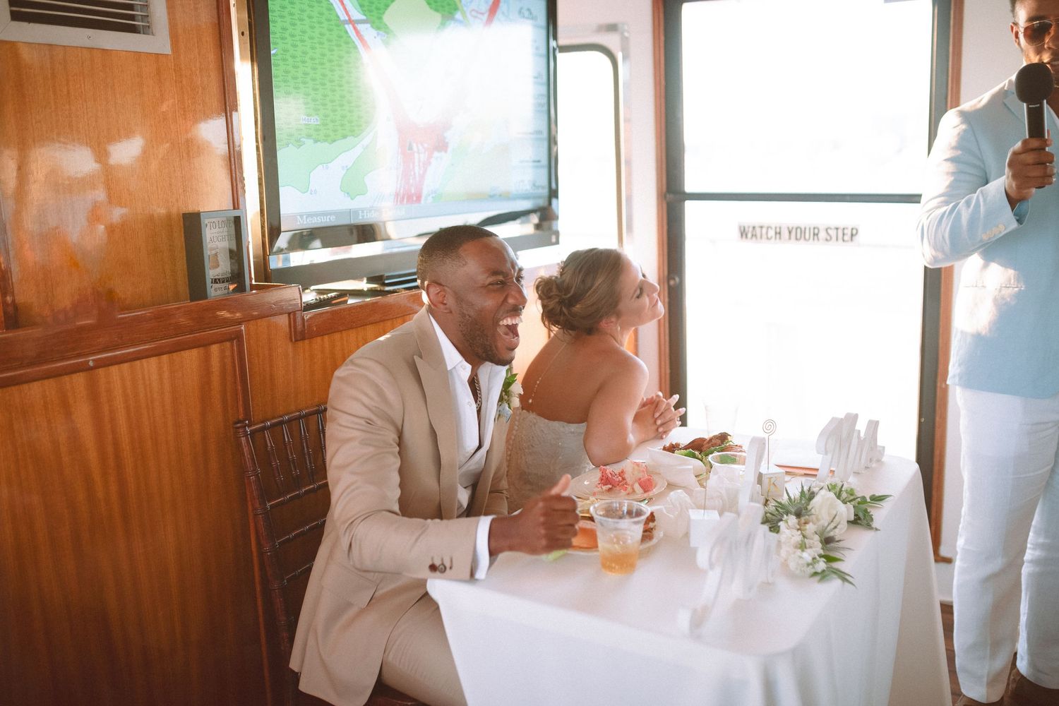 Bride and groom sit at sweetheart table listening to toasts during boat wedding reception.