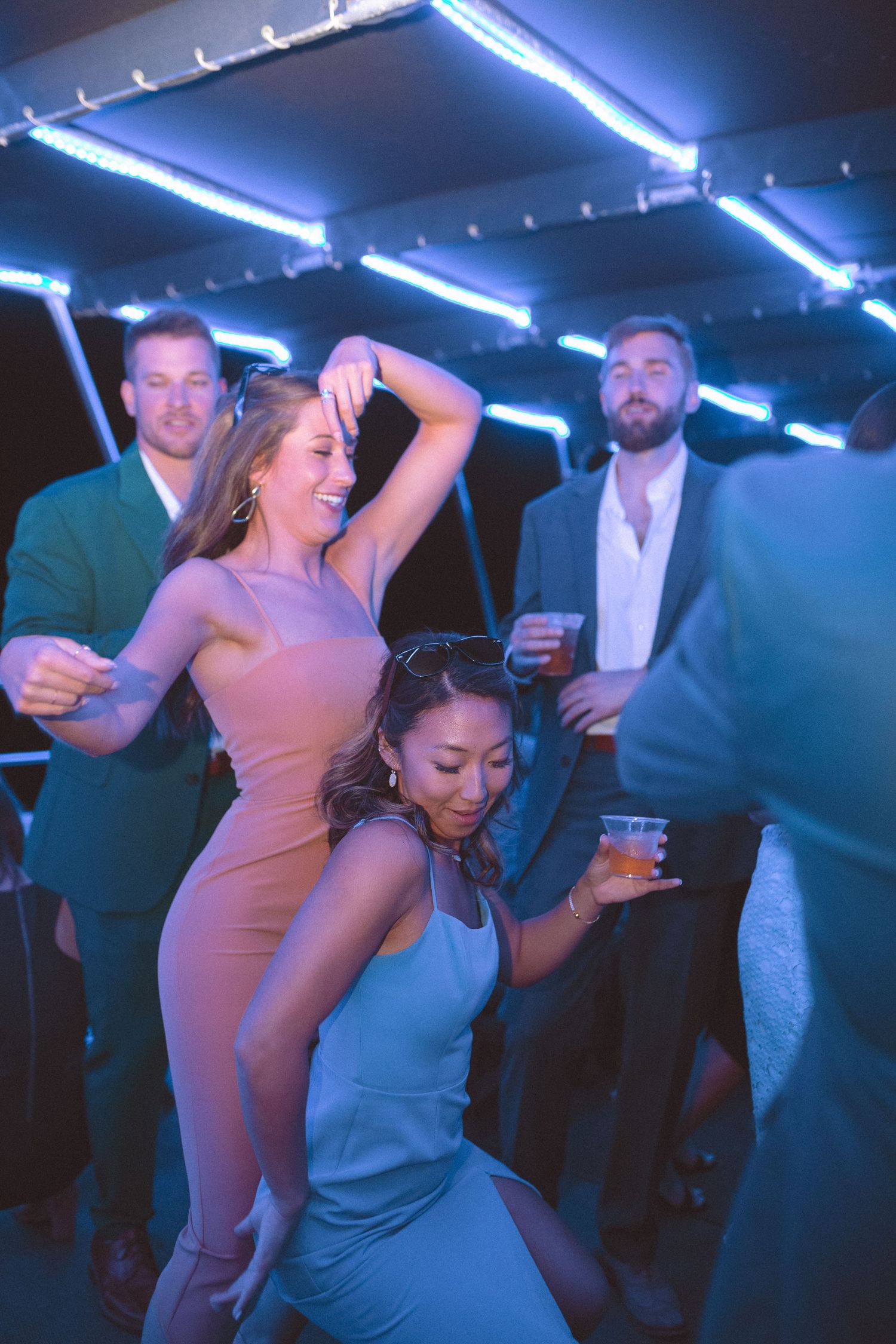 Two women in elegant dresses dancing energetically at a wedding reception with blue neon lighting.