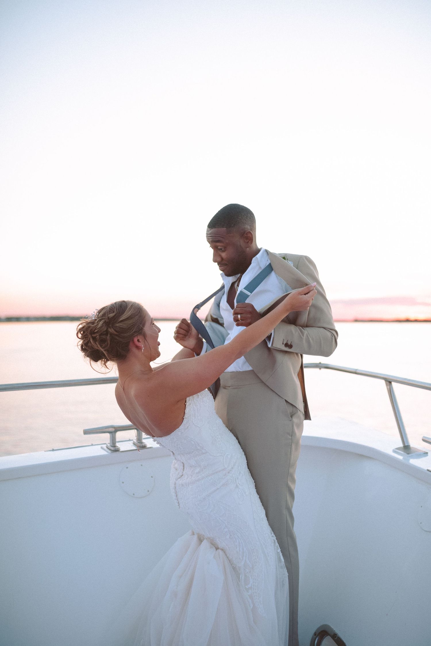 A bride in white lace adjusts her groom's suit jacket on a yacht deck at sunset.