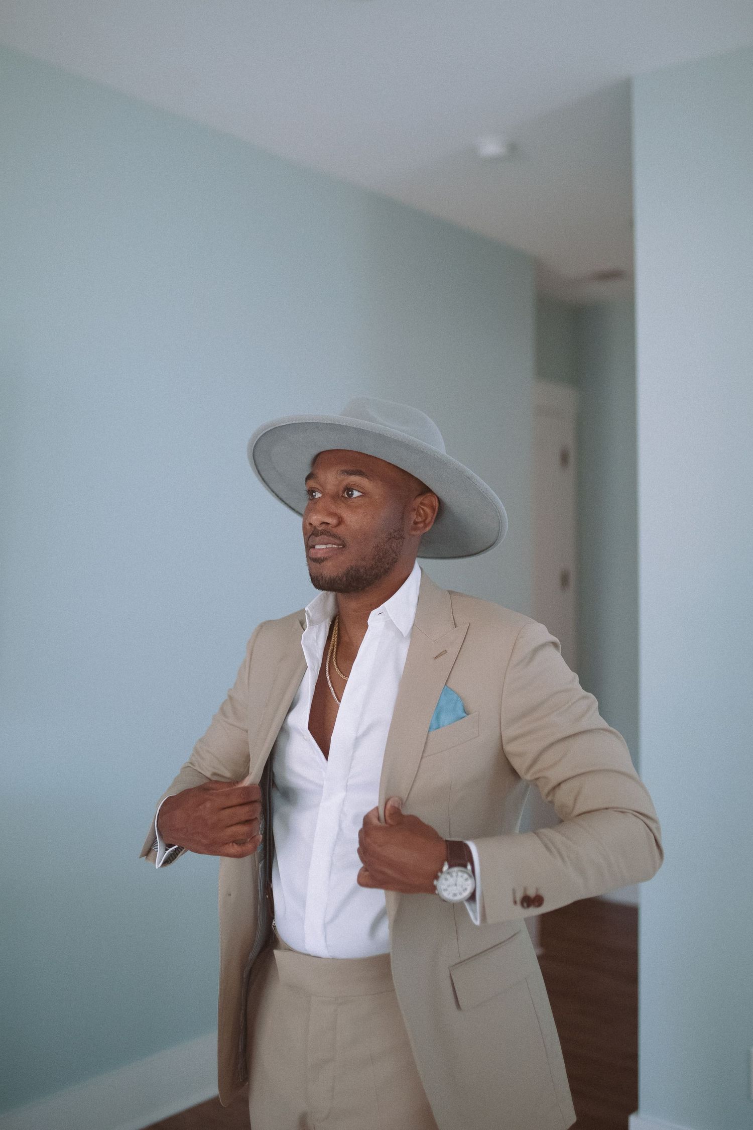 Dapper man in tan suit adjusting jacket lapels wearing gray wide-brim hat and blue pocket square indoors.