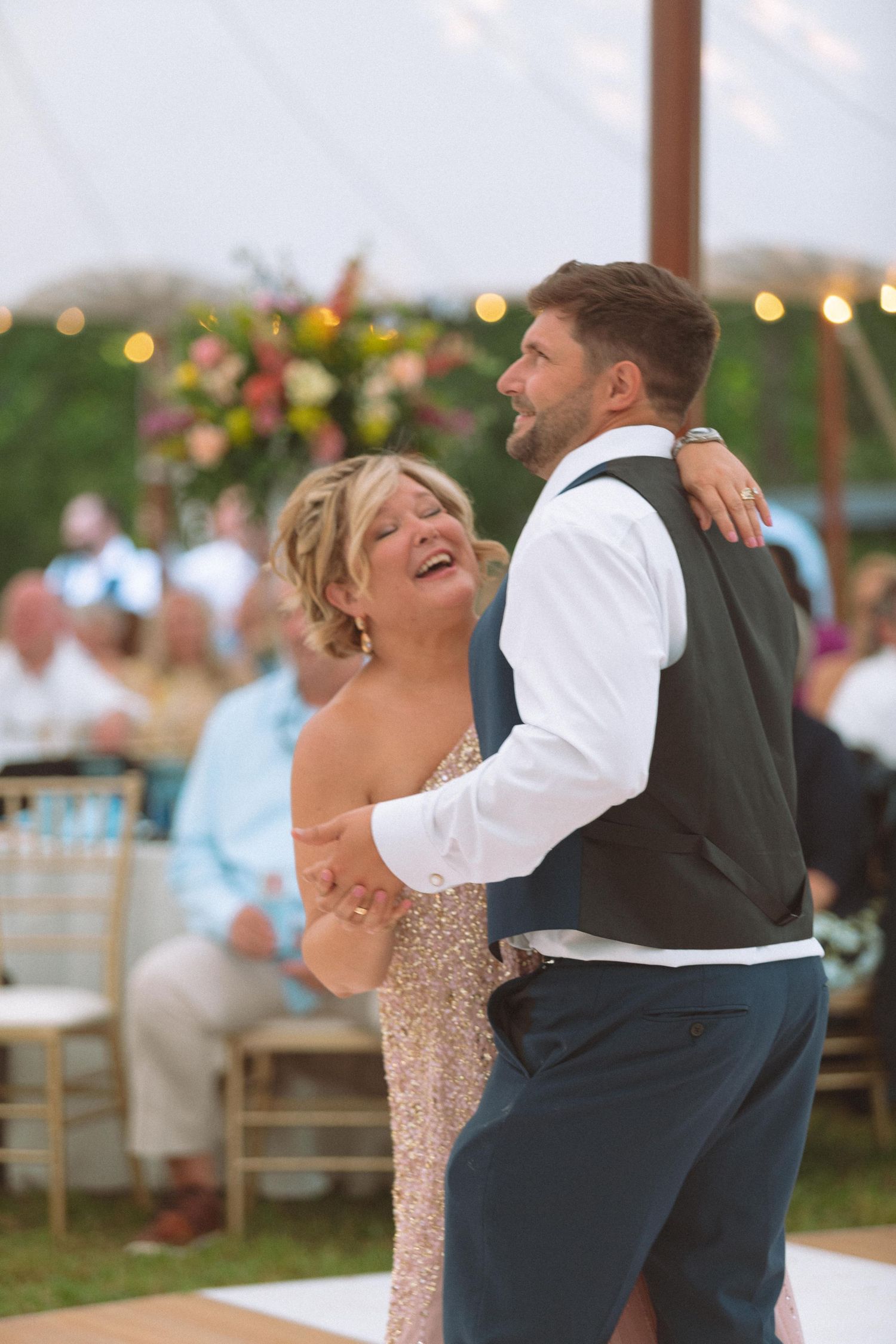 Groom dancing joyfully with his smiling mother under string lights at an outdoor wedding reception.