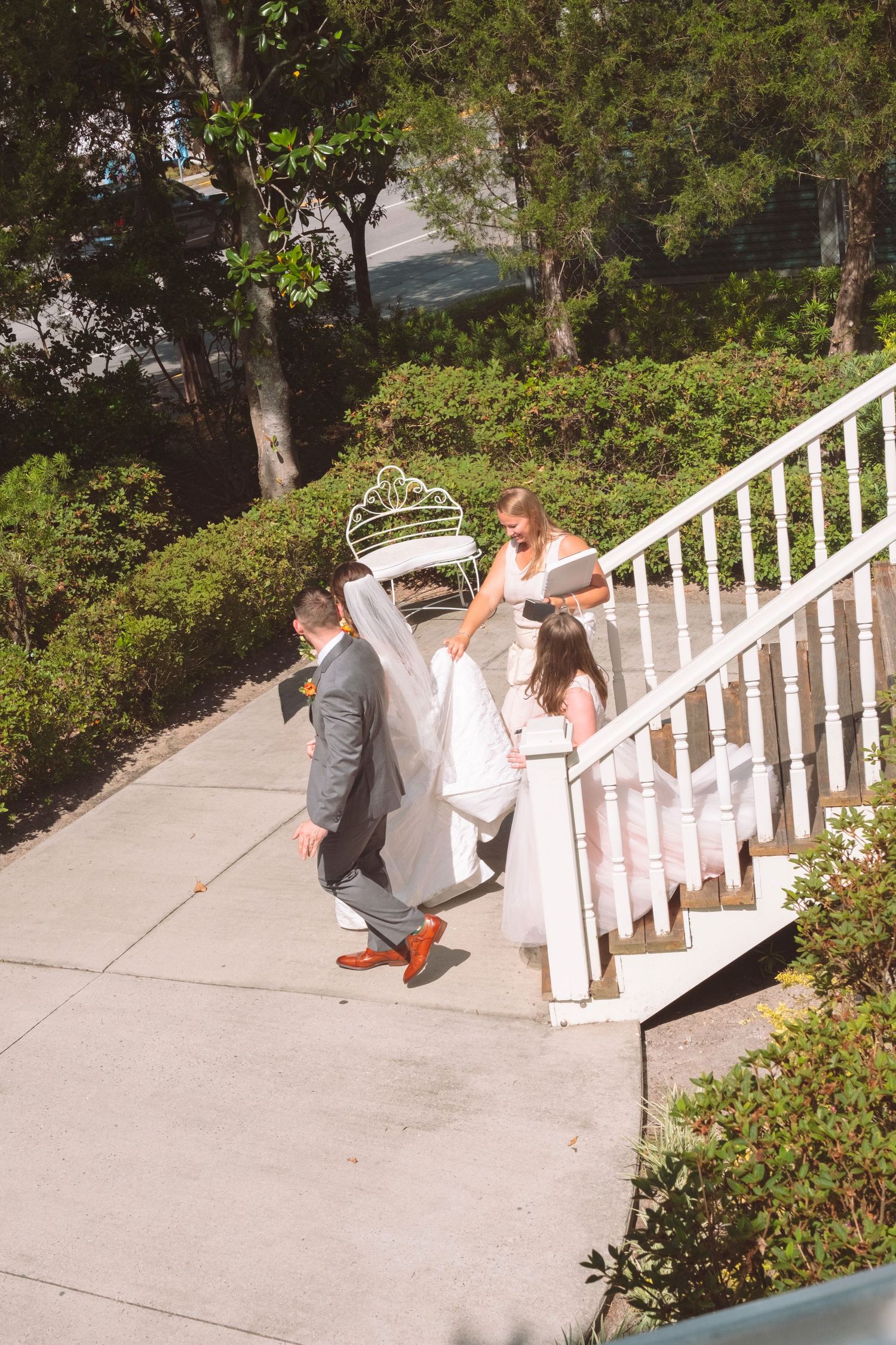 Bride and groom walking up outdoor stairs with wedding dress train being carried by bridesmaid on sunny day.