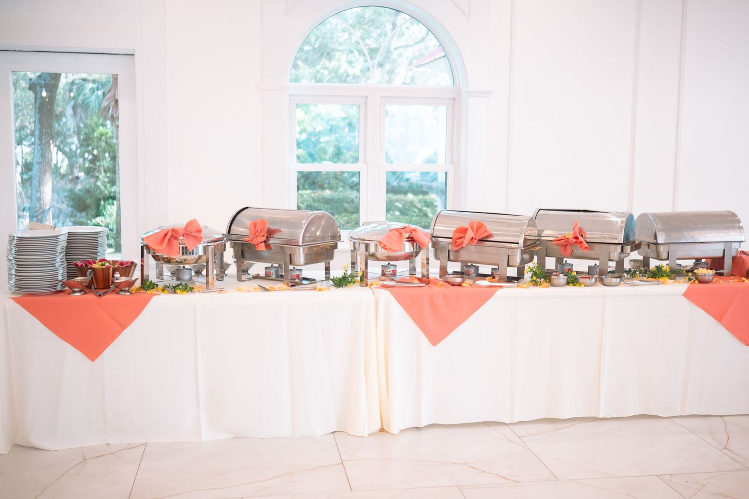 Elegant buffet table with chafing dishes on coral linen runners in a bright white reception hall.