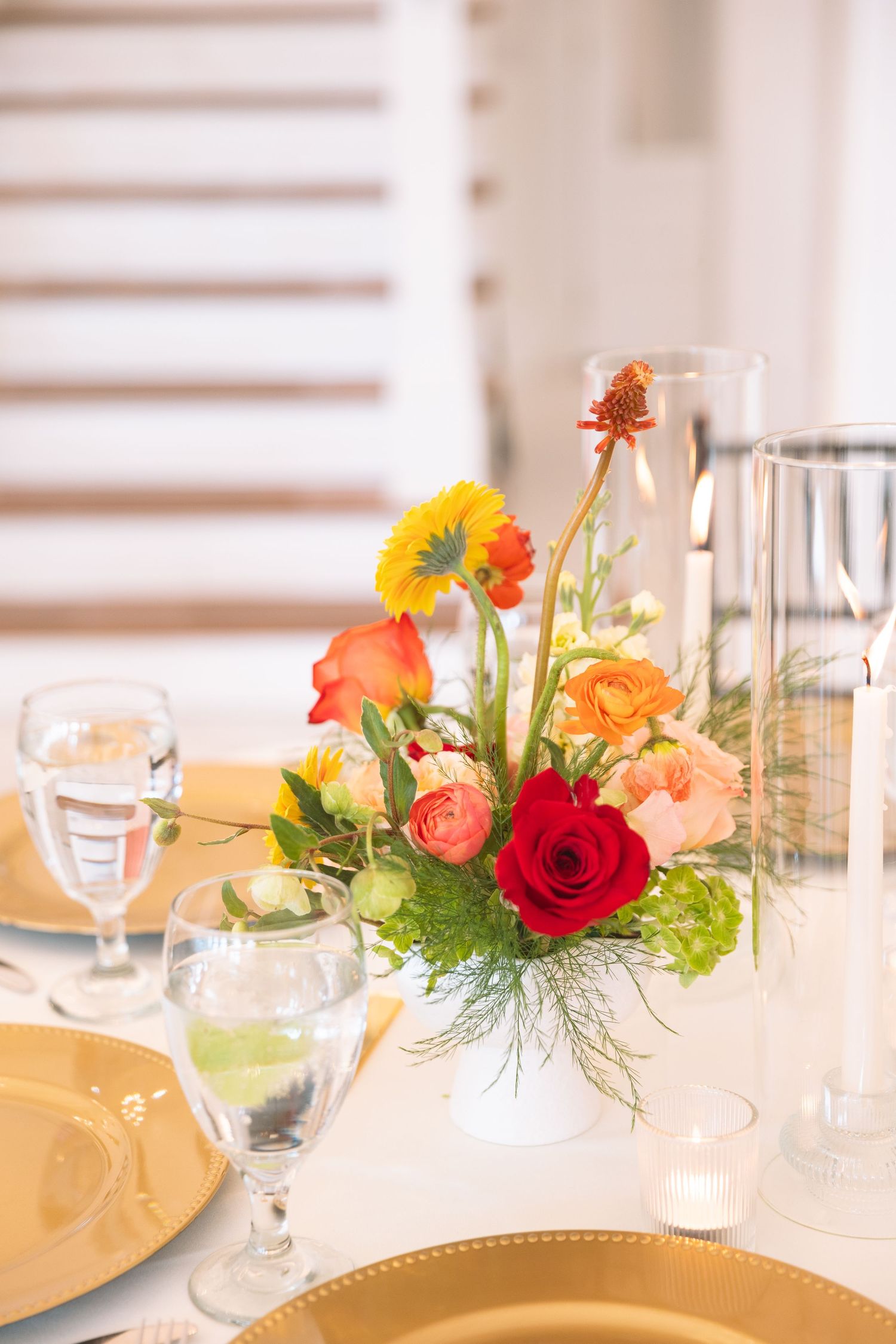 Elegant wedding table centerpiece with red roses, orange gerberas, and peach blooms in white vase with gold charger plates.