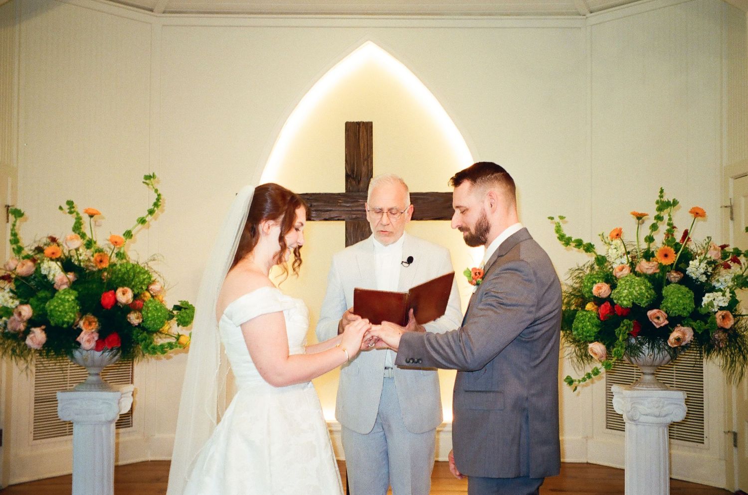 Bride and groom exchange vows at church altar with cross backdrop and floral arrangements.