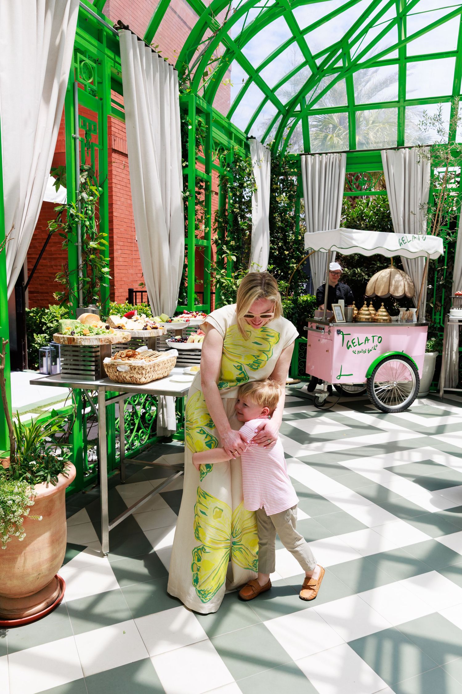 A mother in a floral dress hugs her toddler near a dessert table in a bright green garden pavilion.