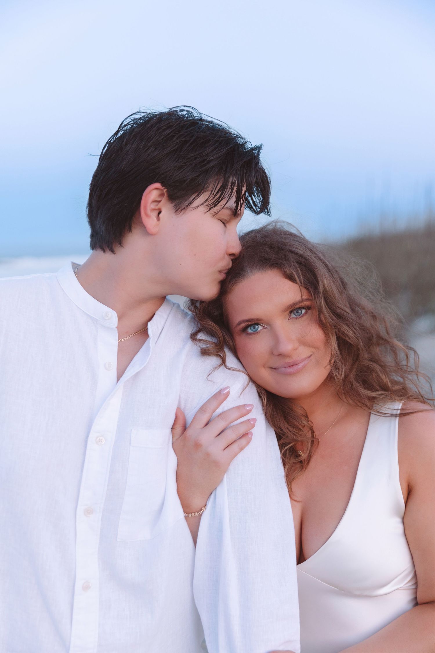 A romantic couple embraces on a beach, the woman smiling with blue eyes as the man whispers in her ear.