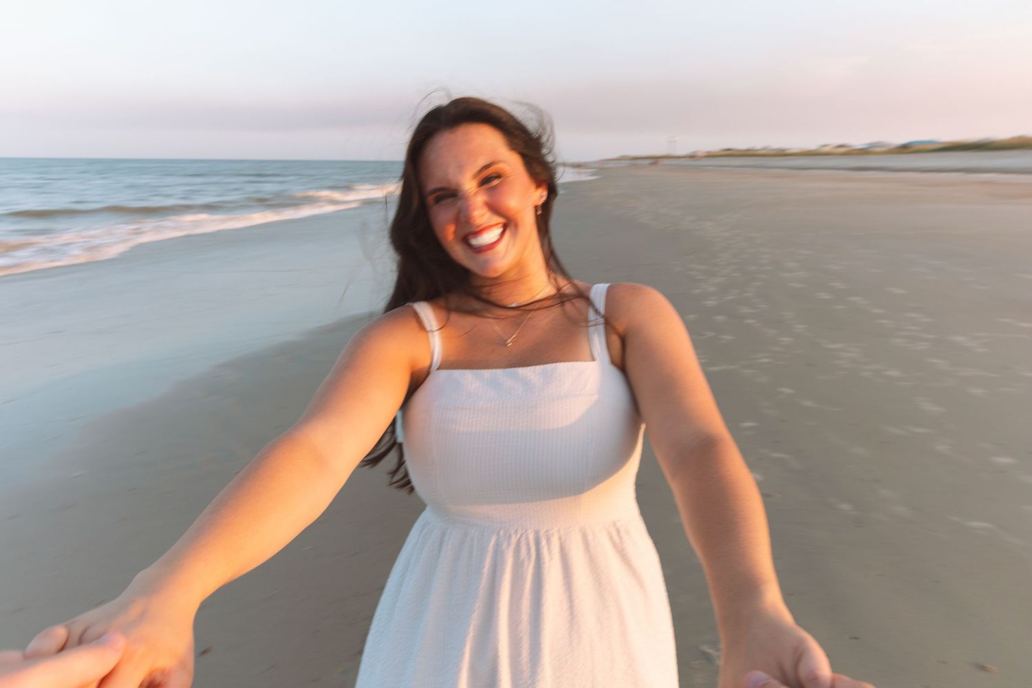 Smiling woman in white sundress holding hands on a sandy beach at golden hour sunset.