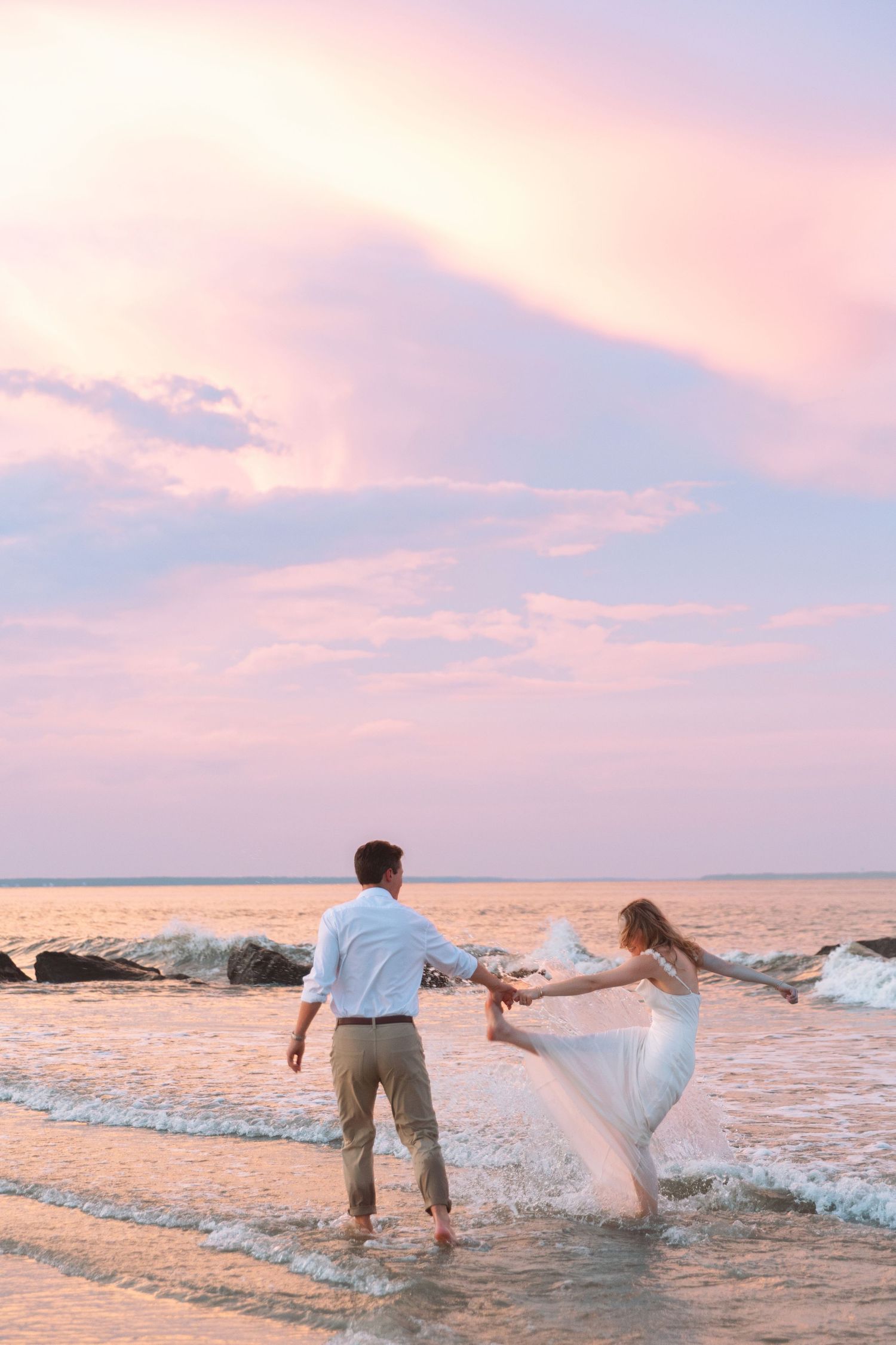 Couple holding hands dancing in ocean waves at sunset with pink and purple sky behind them.