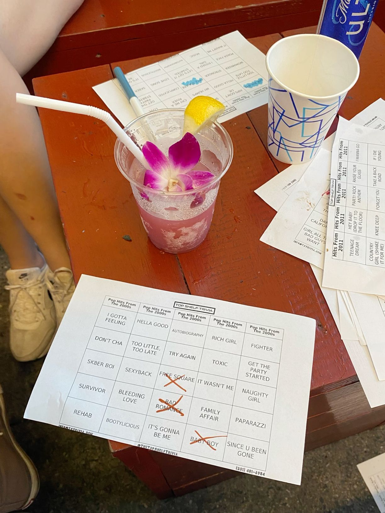 Pink cocktail with orchid garnish beside a Bingo card with crossed-off songs at a music event.