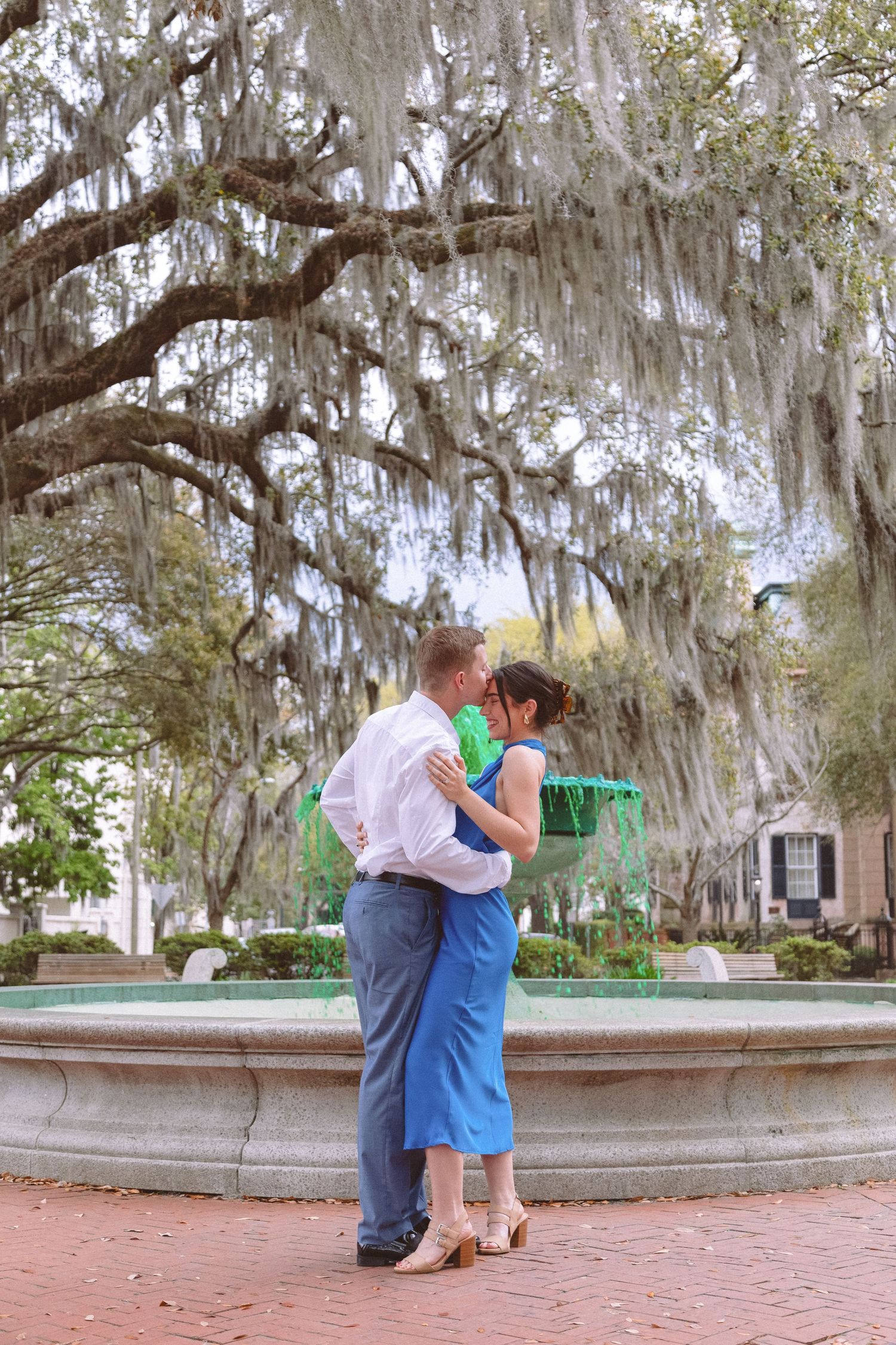 Couple sharing a romantic kiss by a fountain under Spanish moss-draped oak trees in a park.