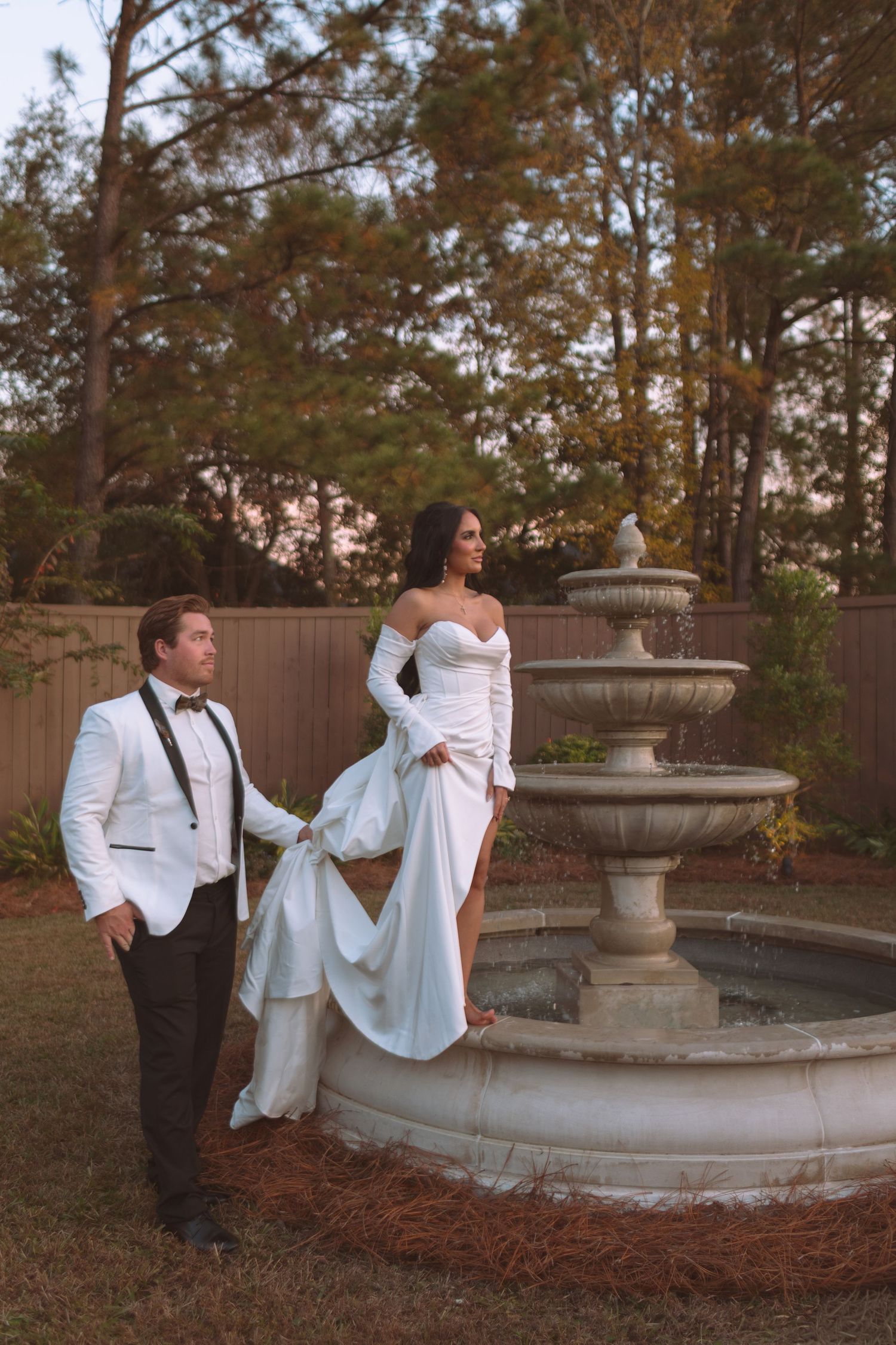 Bride in white off-shoulder gown stands atop a stone fountain while groom in white tuxedo holds her dress outdoors.