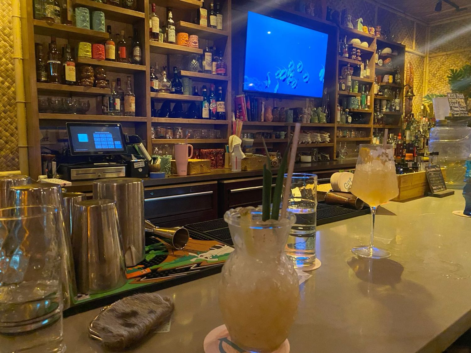 Craft cocktails and mixing pitcher on bar counter with well-stocked backbar and TV screen.