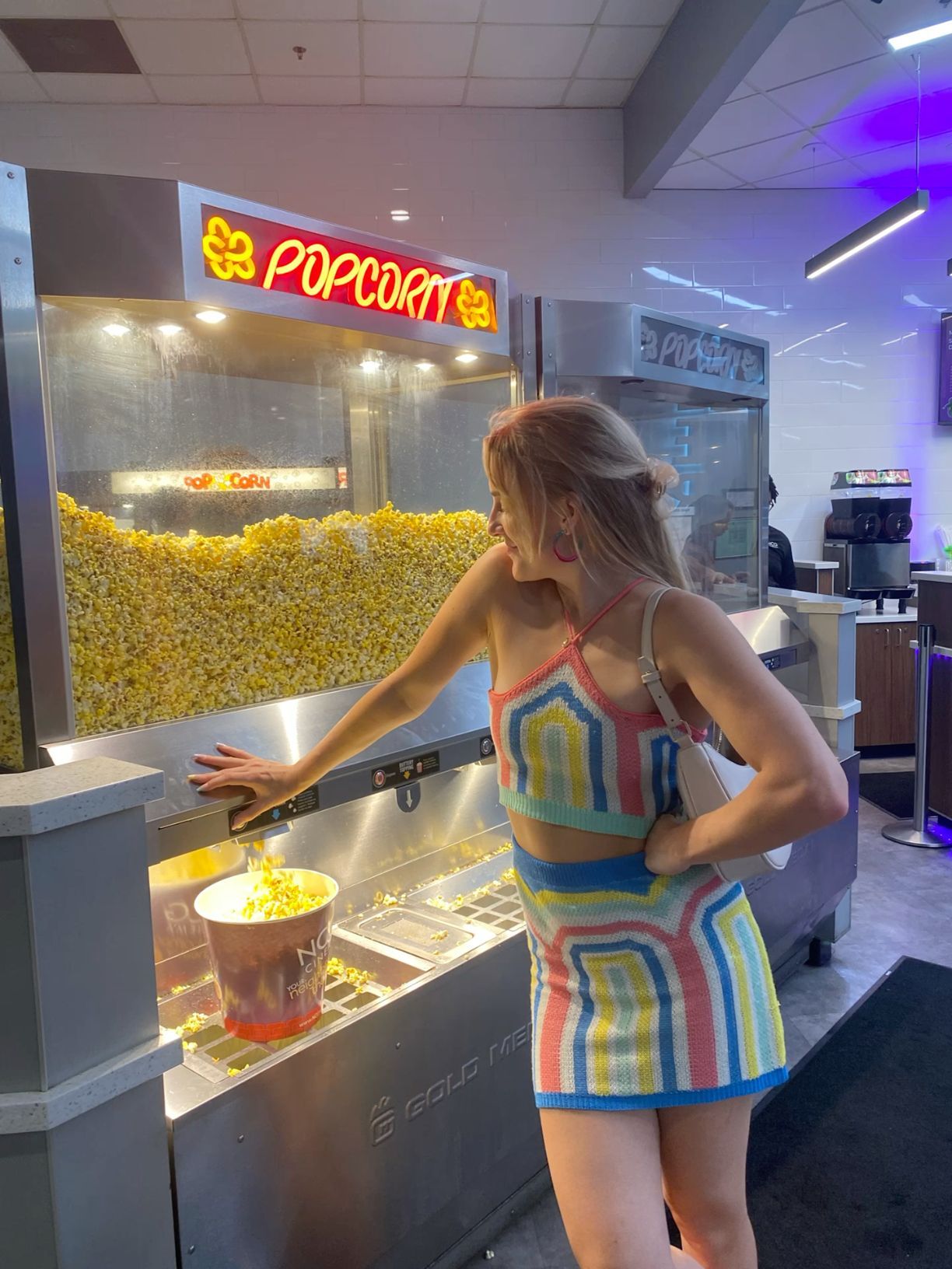 Blonde woman in colorful striped outfit scooping popcorn at a bright cinema concession stand.