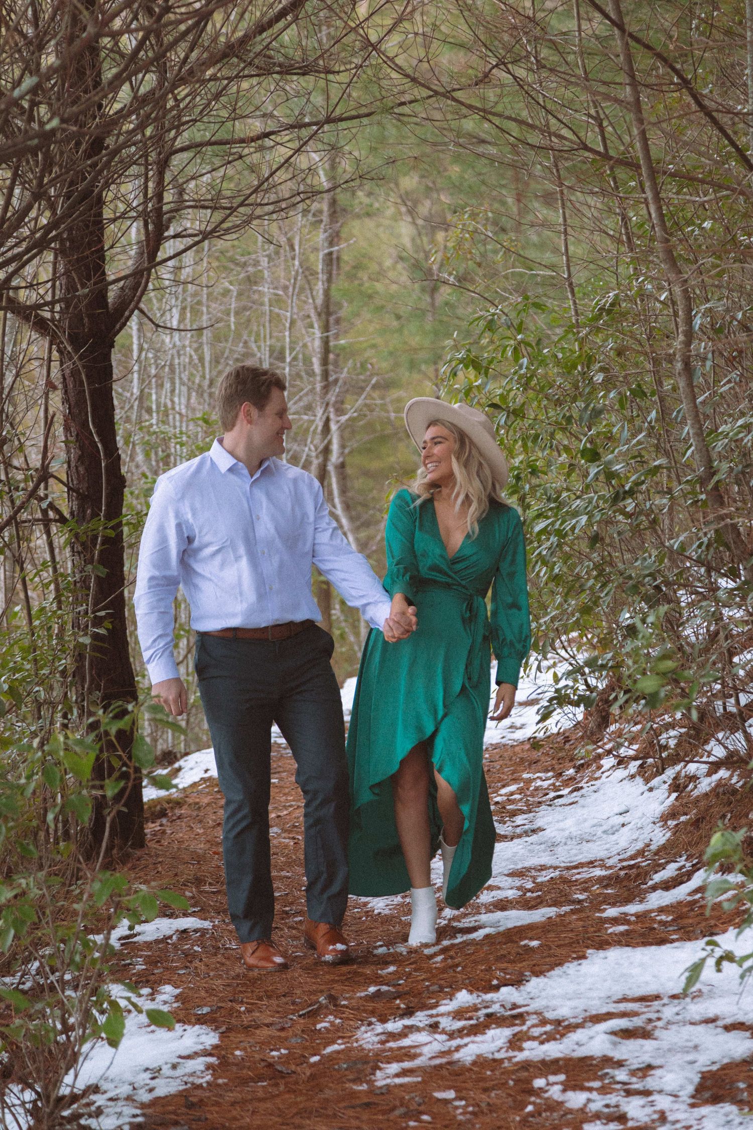Couple walking hand in hand along a snowy forest path, smiling at each other in winter woods.