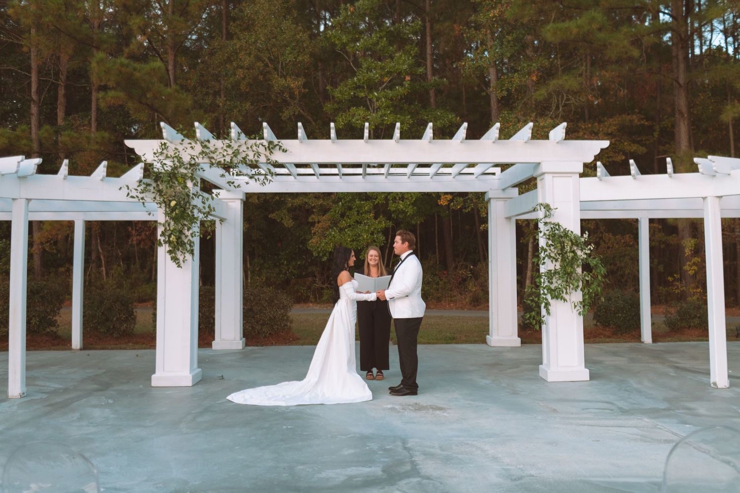 Bride and groom exchange vows under a white pergola adorned with greenery at an outdoor wedding ceremony.