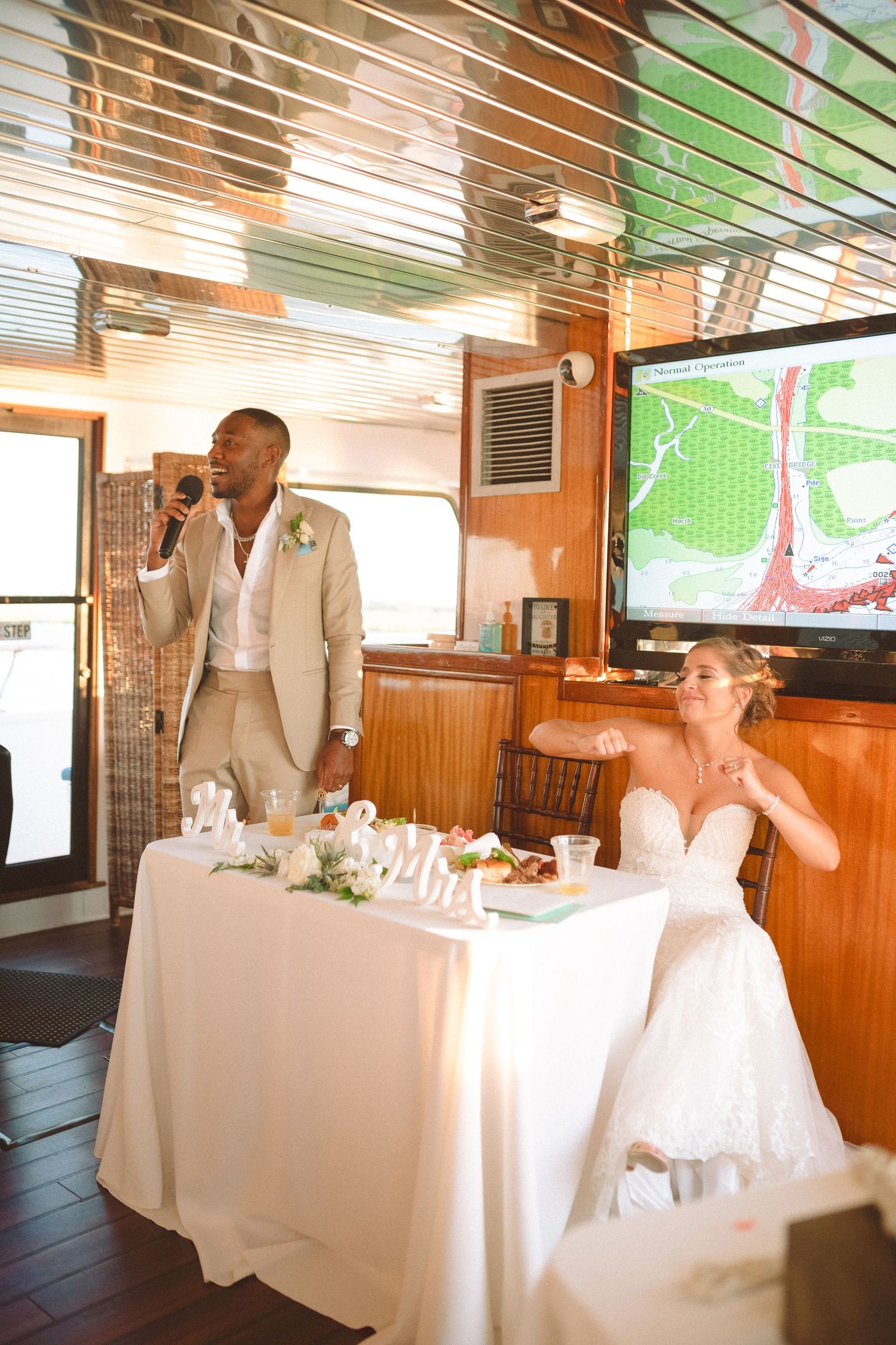 Groom speaks into microphone at sweetheart table while bride laughs during boat wedding reception.