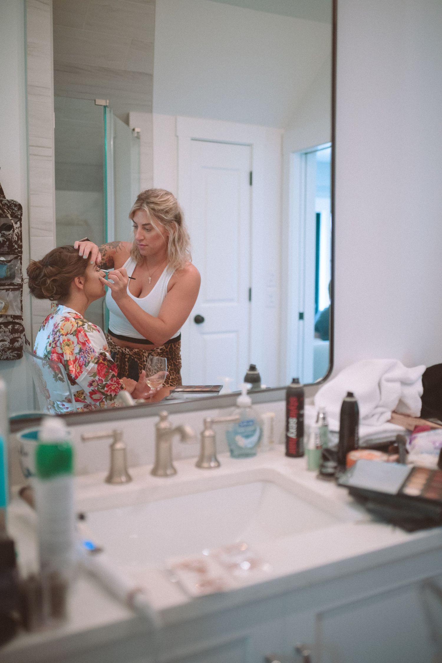 Makeup artist applying makeup to bride reflected in bathroom mirror surrounded by beauty products.
