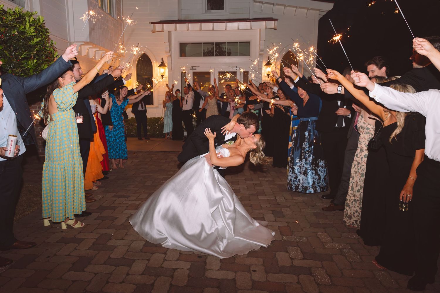 Bride and groom share romantic dip kiss during sparkler send-off exit at elegant outdoor wedding reception at night.