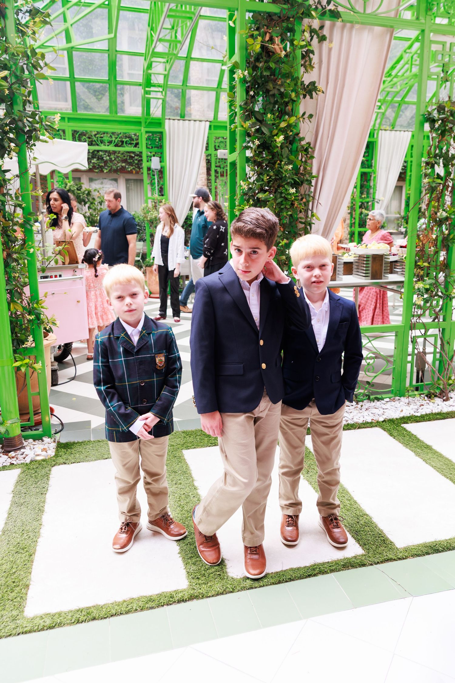 Three well-dressed boys in blazers and khakis pose together at an elegant outdoor event with green arch decor.