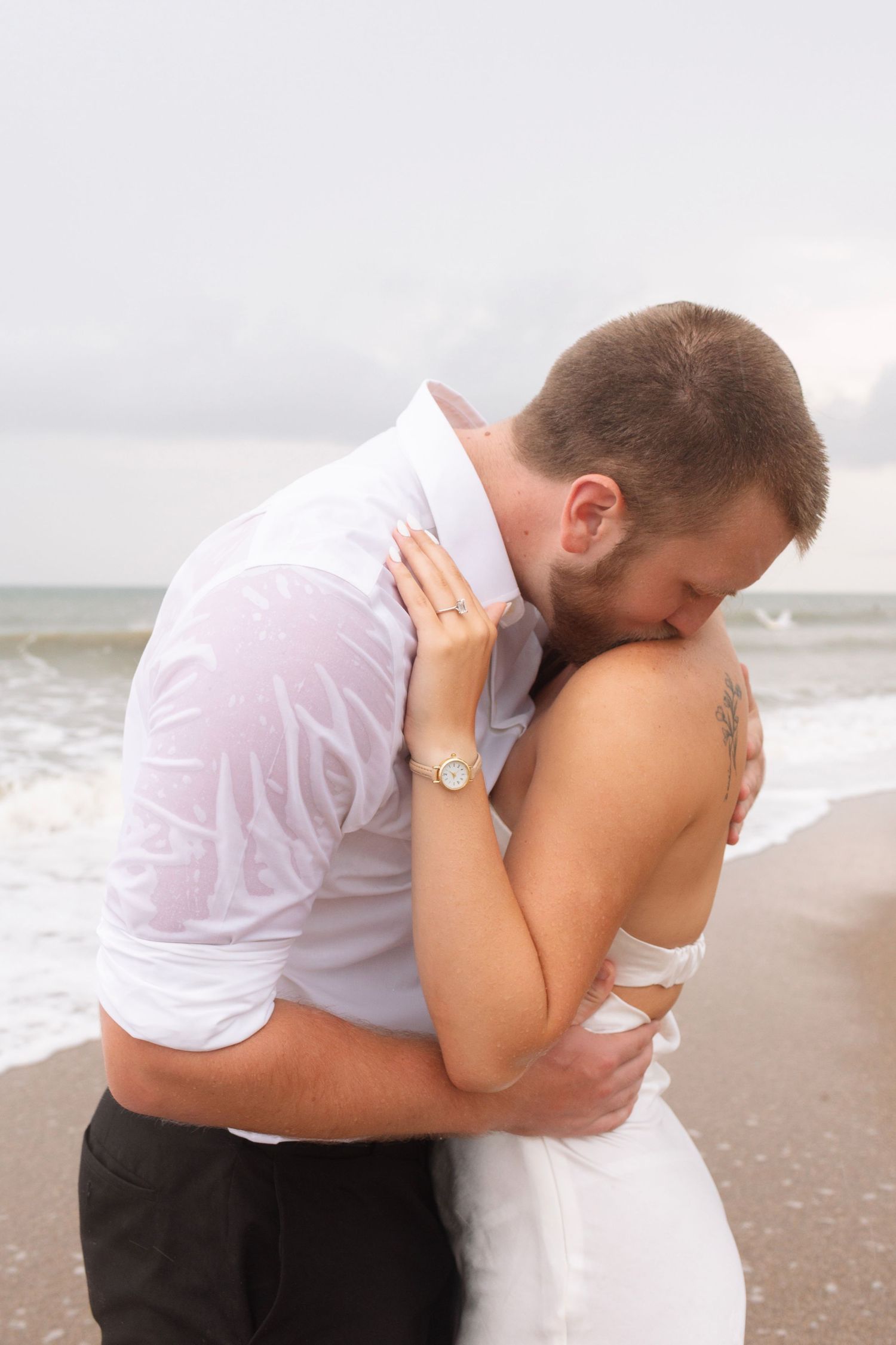 Bride and groom sharing an intimate embrace on a cloudy beach, both dressed in white.