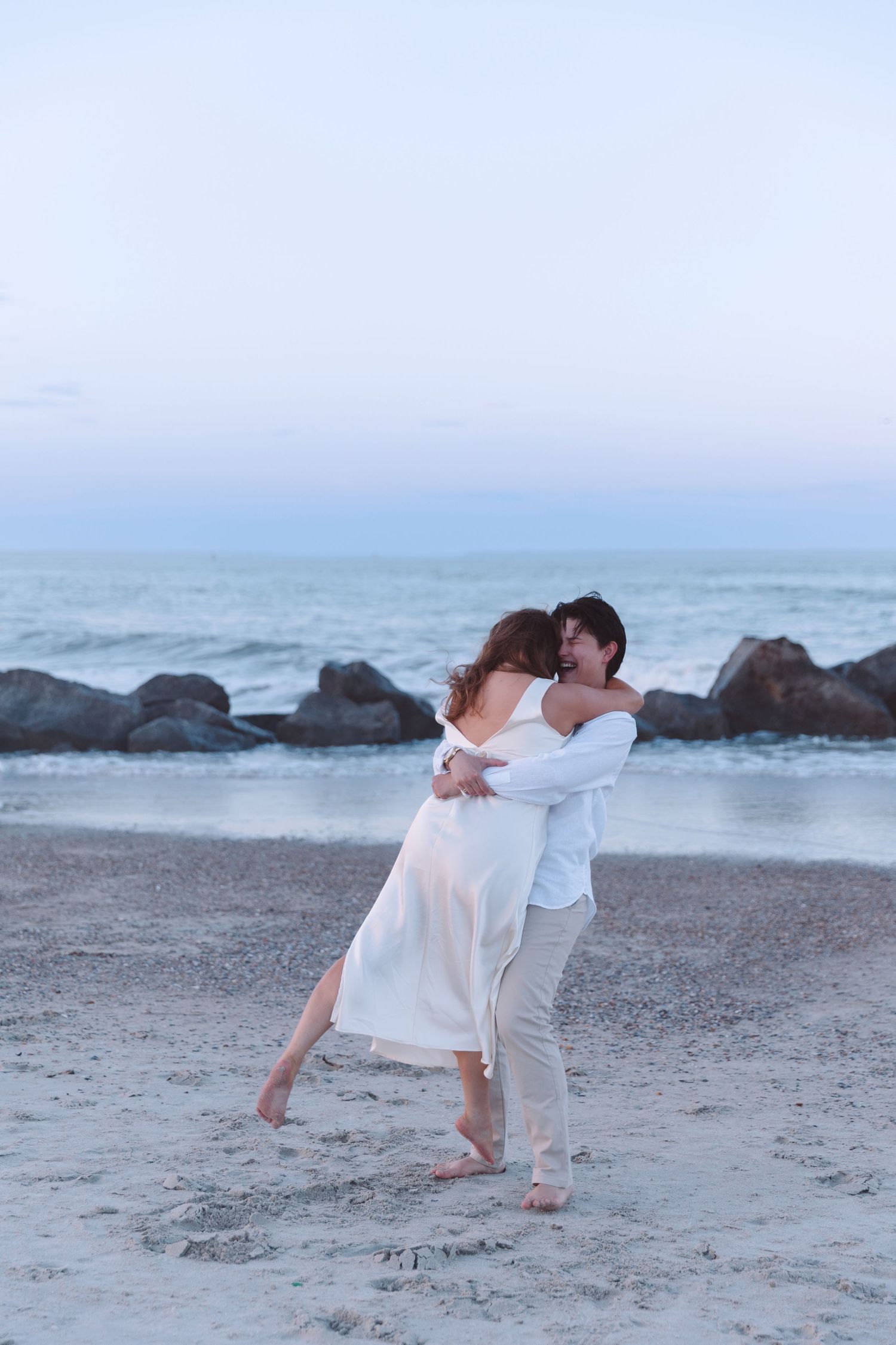 Couple dancing romantically on a sandy beach at dusk with rocky coastline and calm ocean behind them.