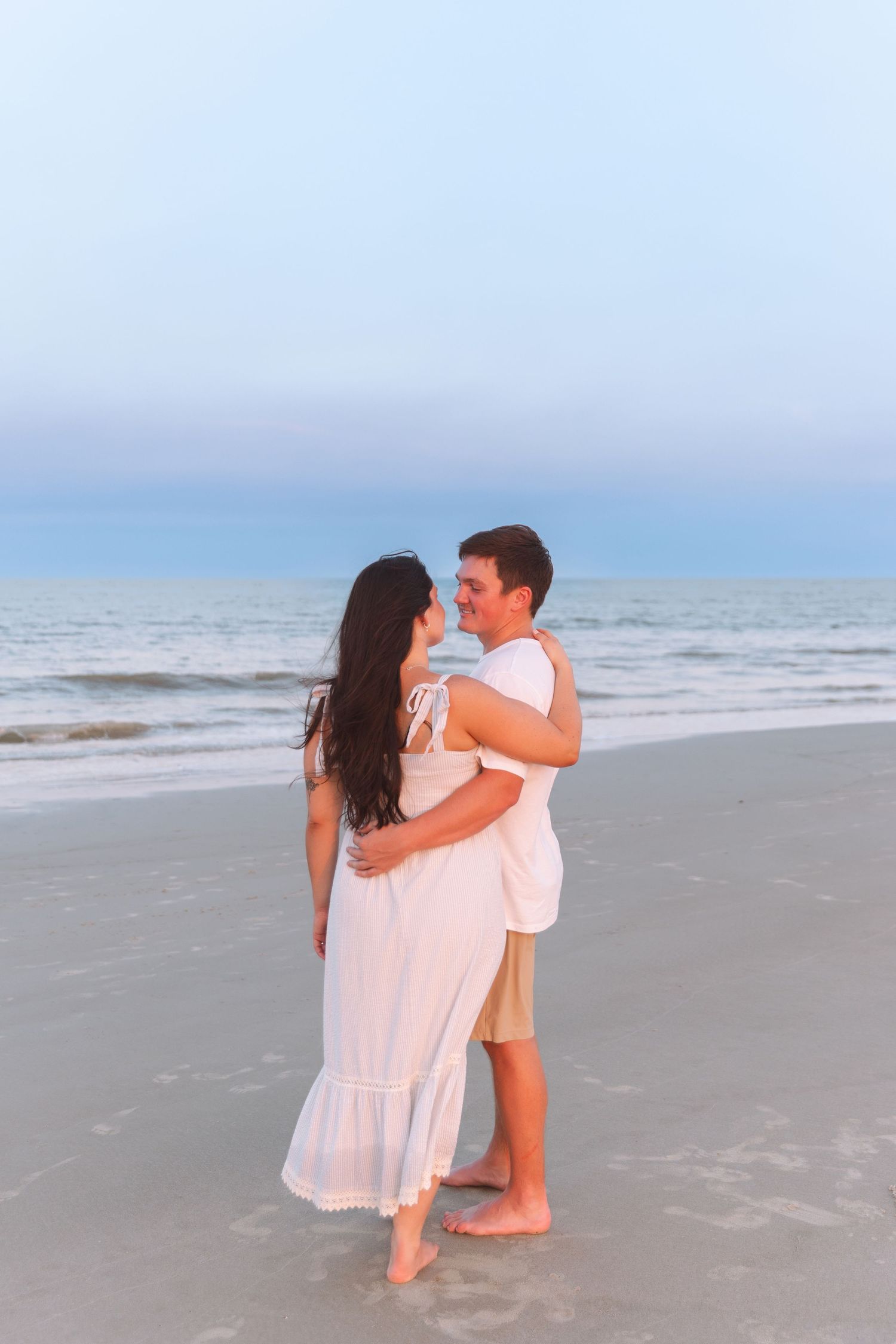 Couple embracing barefoot on a sandy beach at dusk, woman in flowing white maxi dress held by man in white shirt.