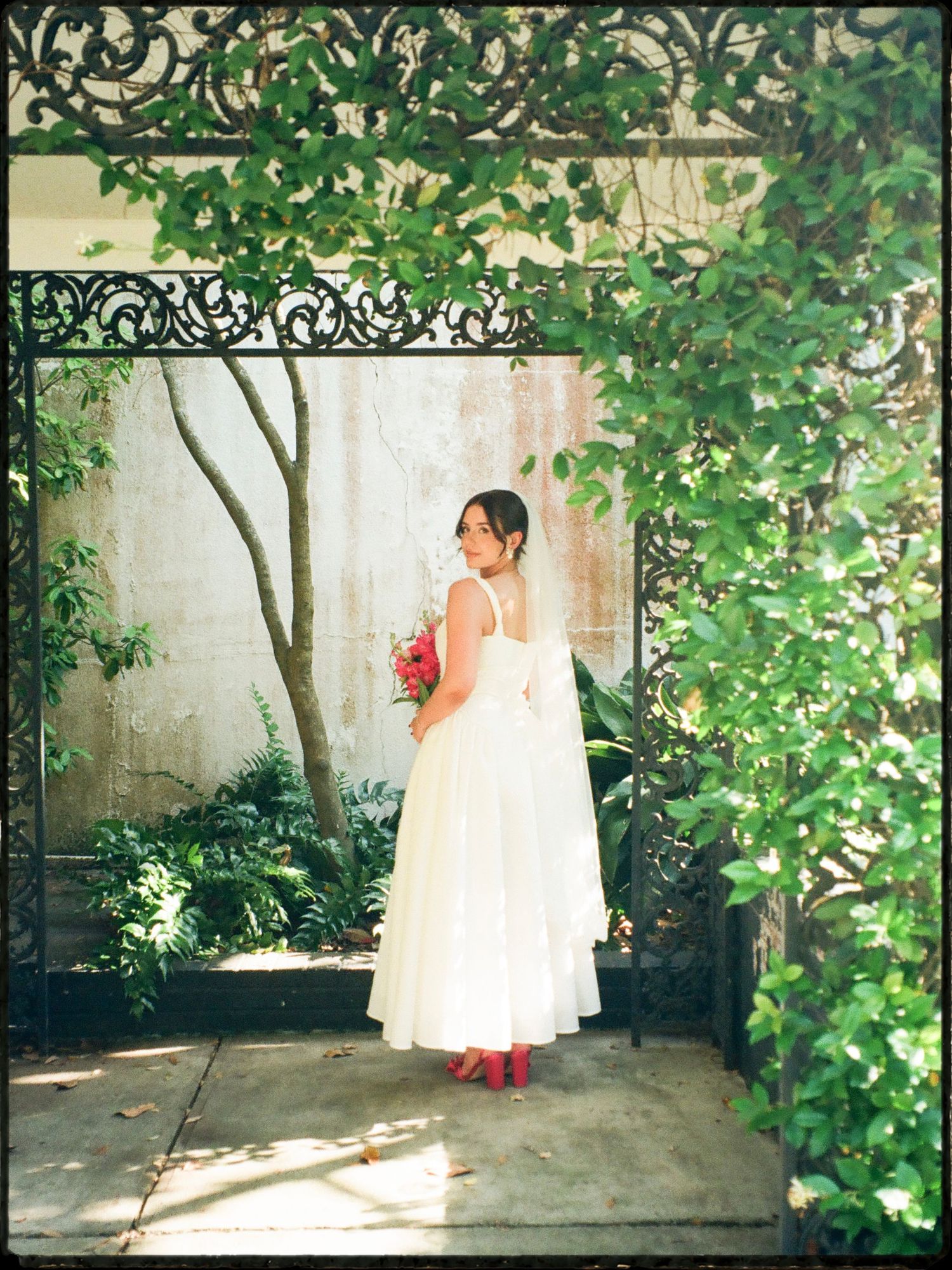 Bride in white tea-length gown with red heels holds pink bouquet under ornate iron garden gate.