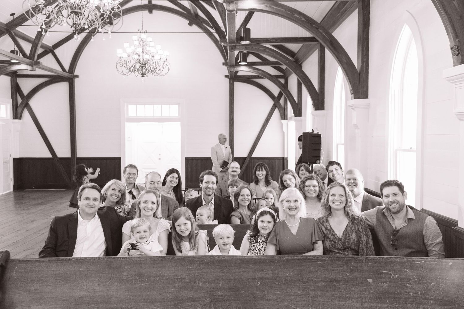 Black and white photo of large wedding guest group posing together inside a rustic chapel venue.