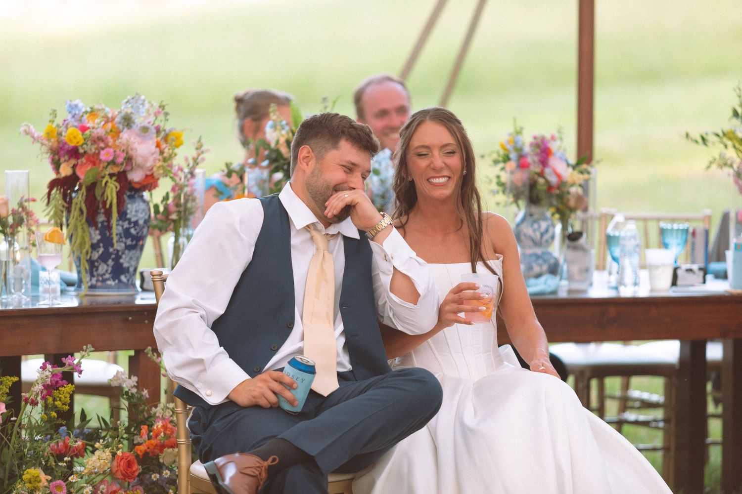 Smiling bride and groom laughing together at sweetheart table during outdoor wedding reception.
