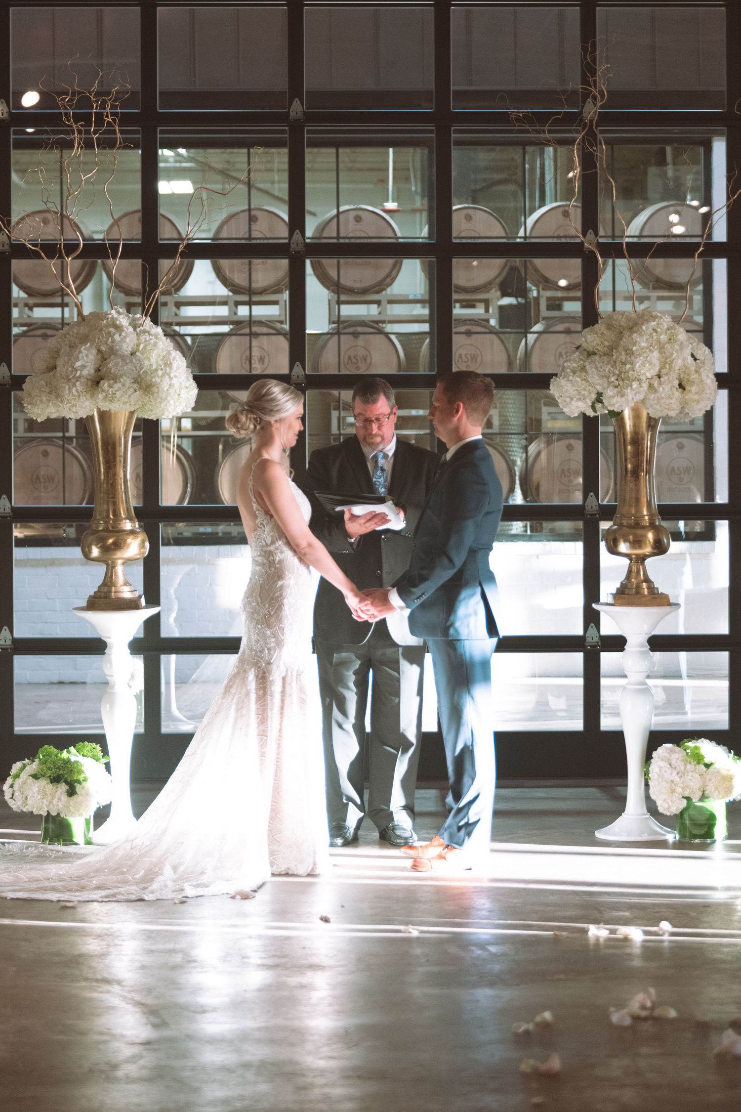 Bride and groom exchange vows before officiant with white floral arrangements at distillery wedding venue.