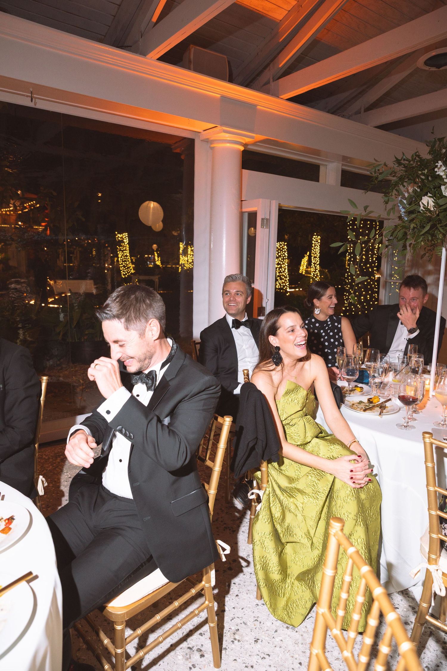 Wedding guests laugh and celebrate at warmly lit evening reception with fairy lights.