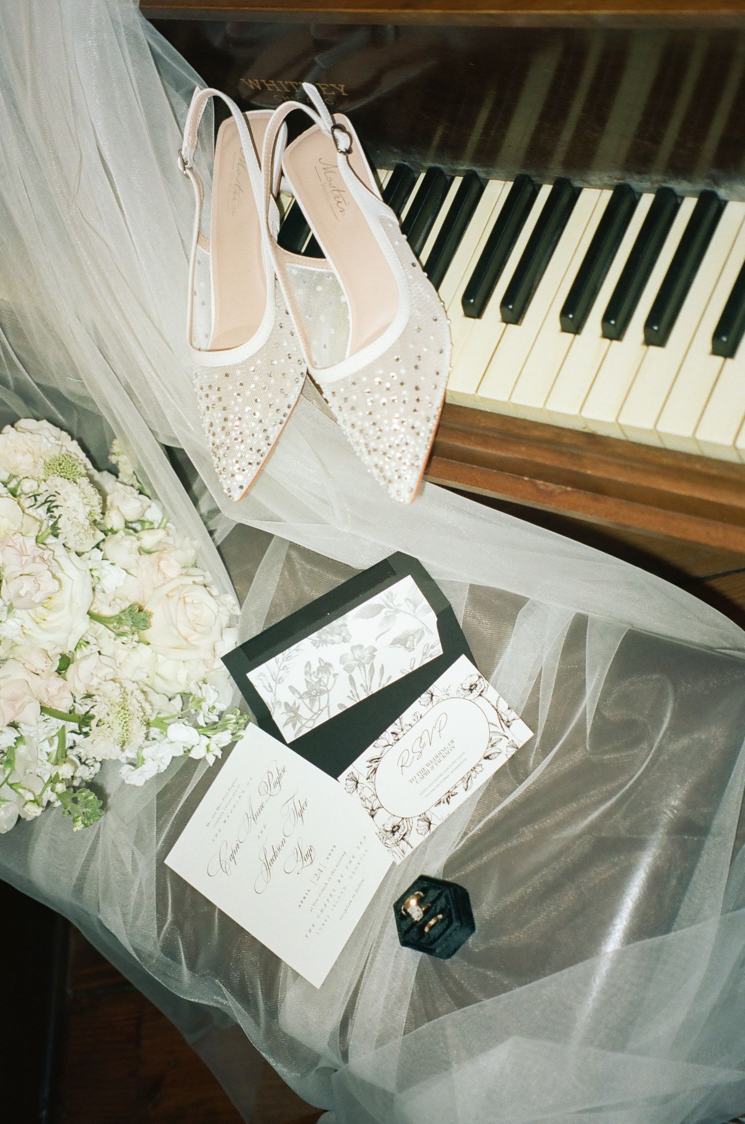 Elegant wedding flat lay featuring crystal-embellished bridal shoes, floral bouquet, invitation suite, and piano keys.