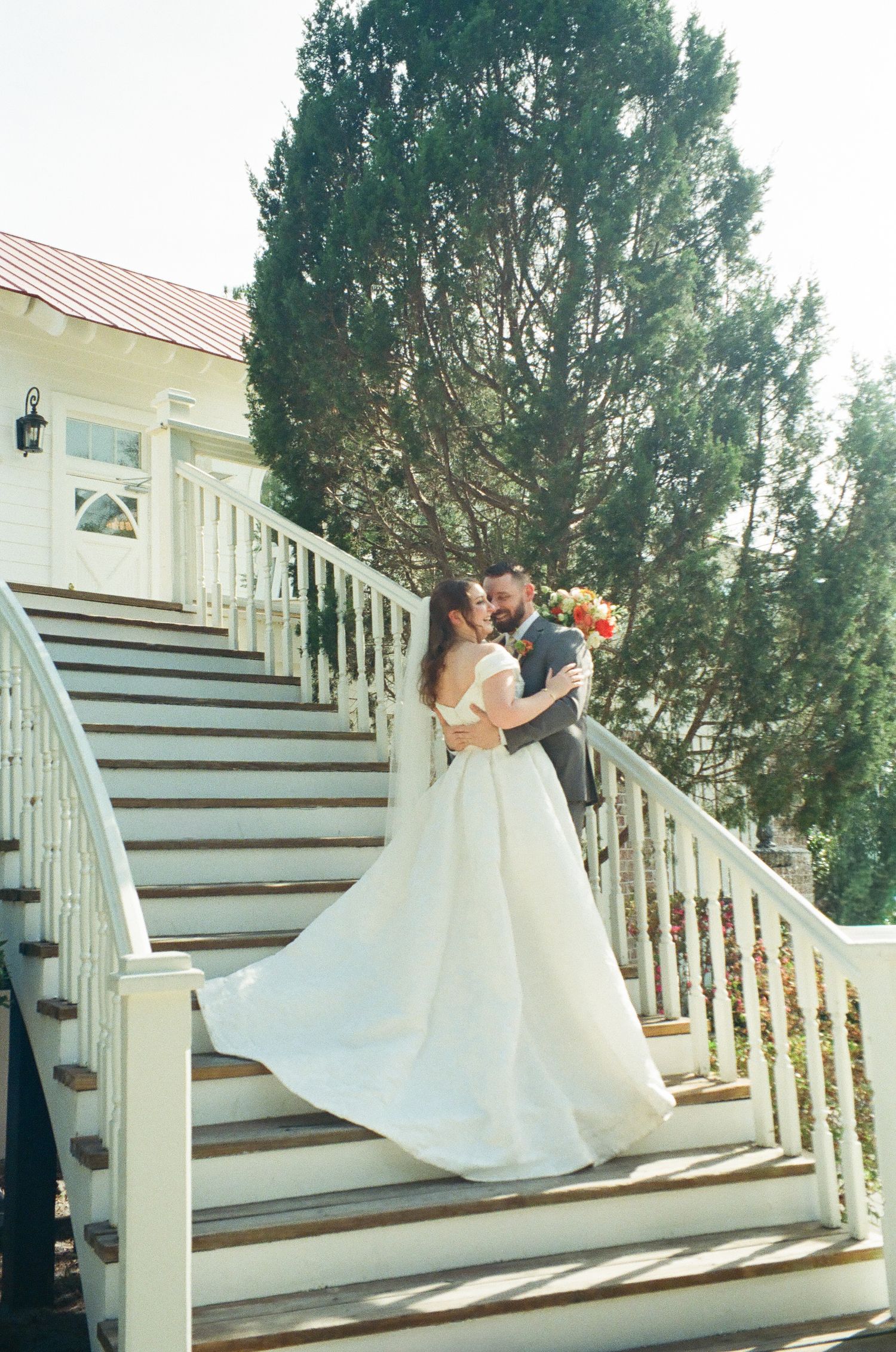 Bride and groom share a romantic embrace on white staircase steps, bride in flowing white gown holding colorful bouquet.