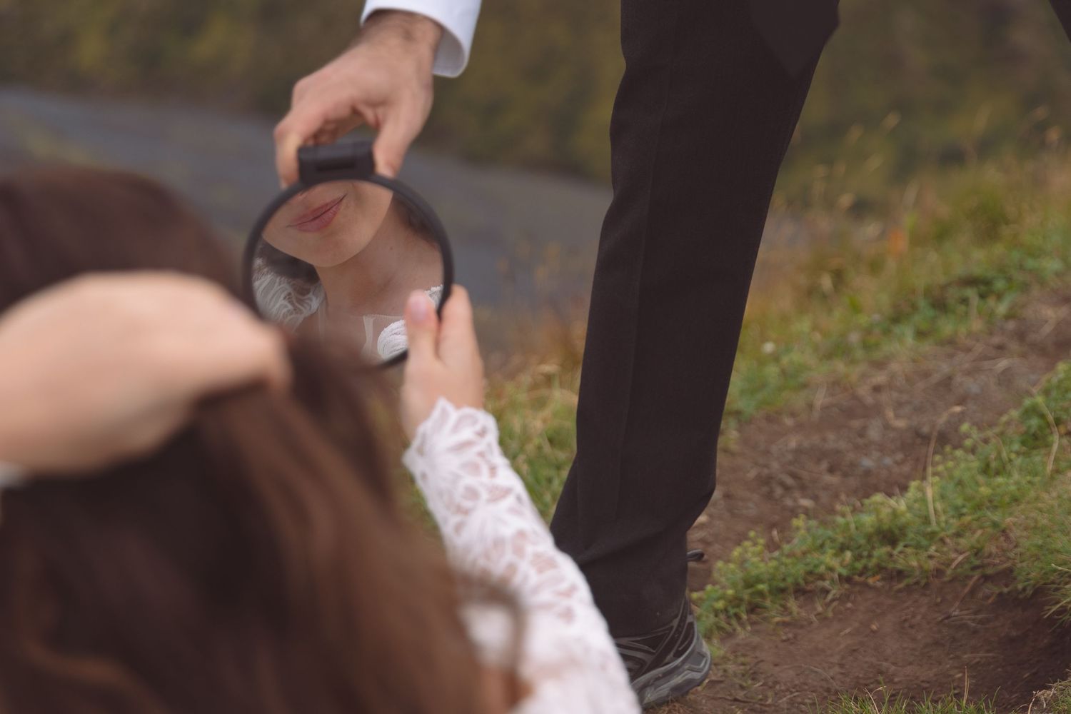 Bride in lace dress holding magnifying glass up to grooms face outdoors on grassy path.