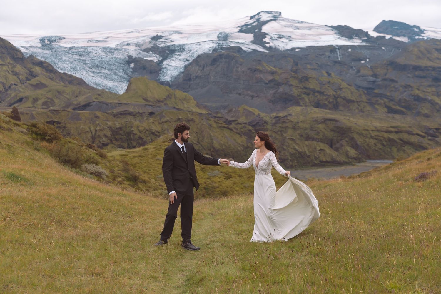 Wedding couple holding hands on green hillside with stunning Icelandic glacier behind them.