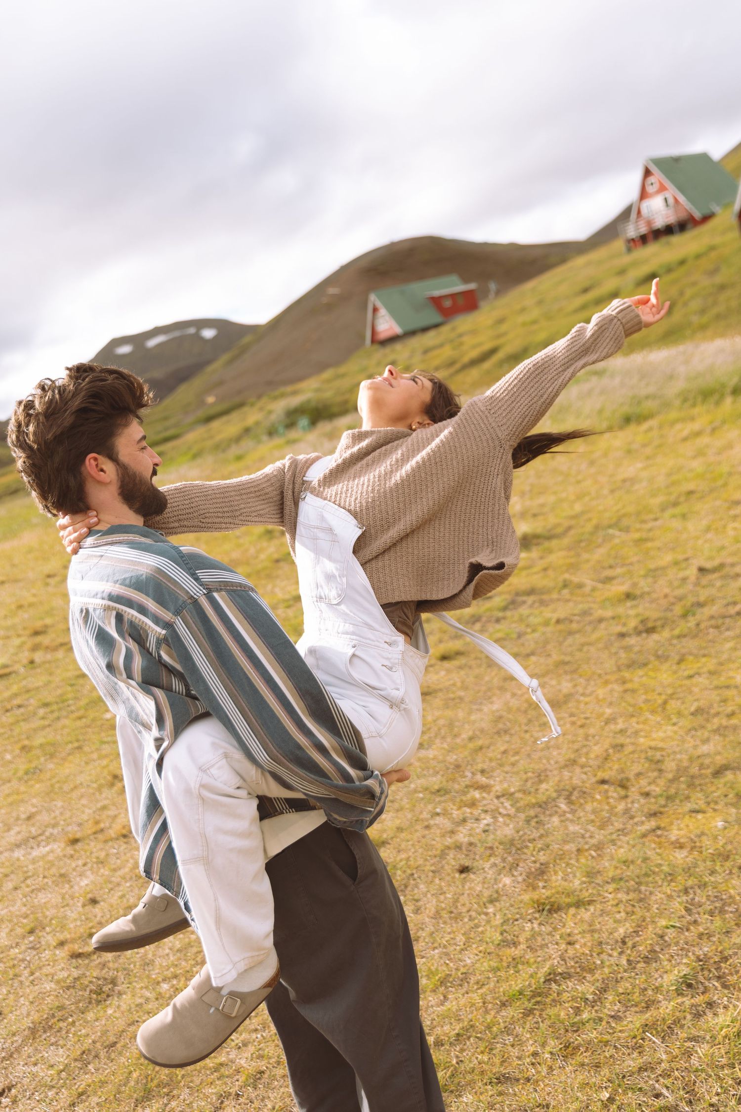 Man carrying woman in his arms on a green hillside in Iceland, she spreads her arms wide with joy.
