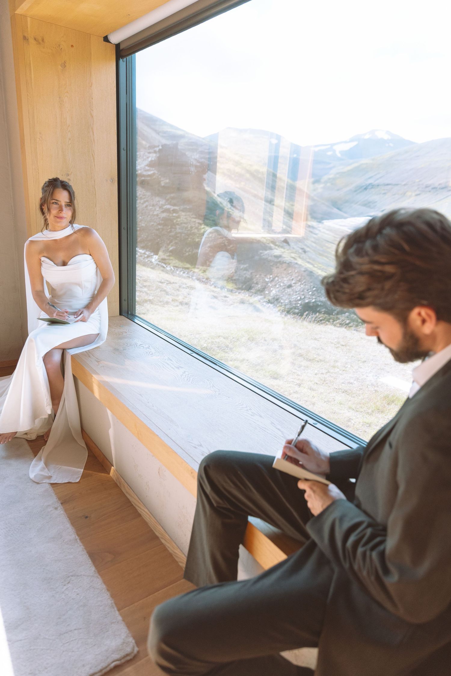 Bride watches groom write wedding vows while seated by panoramic mountain view window.