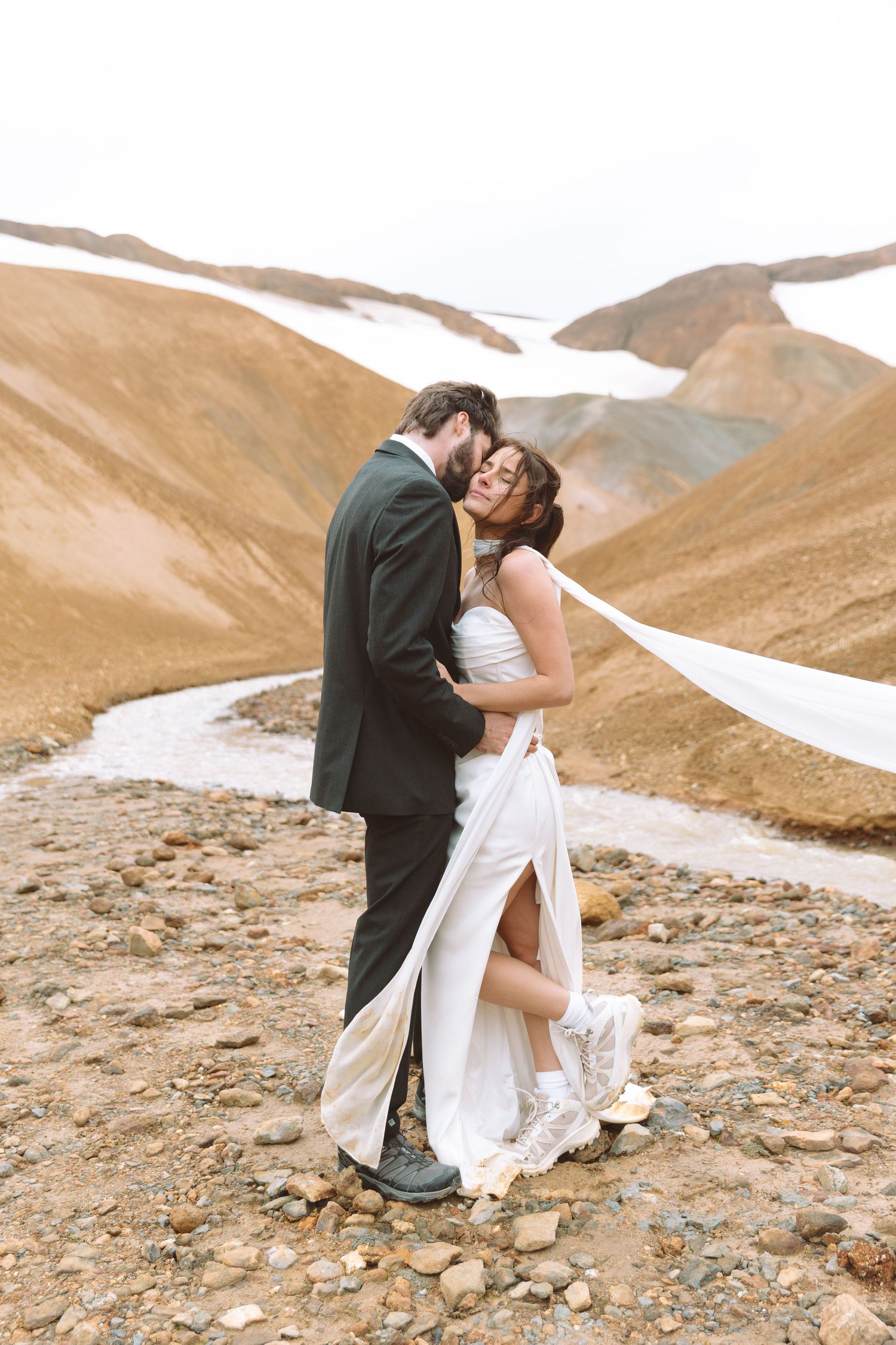 Bride and groom share a close embrace on rocky ground with snow-capped brown mountains behind them.