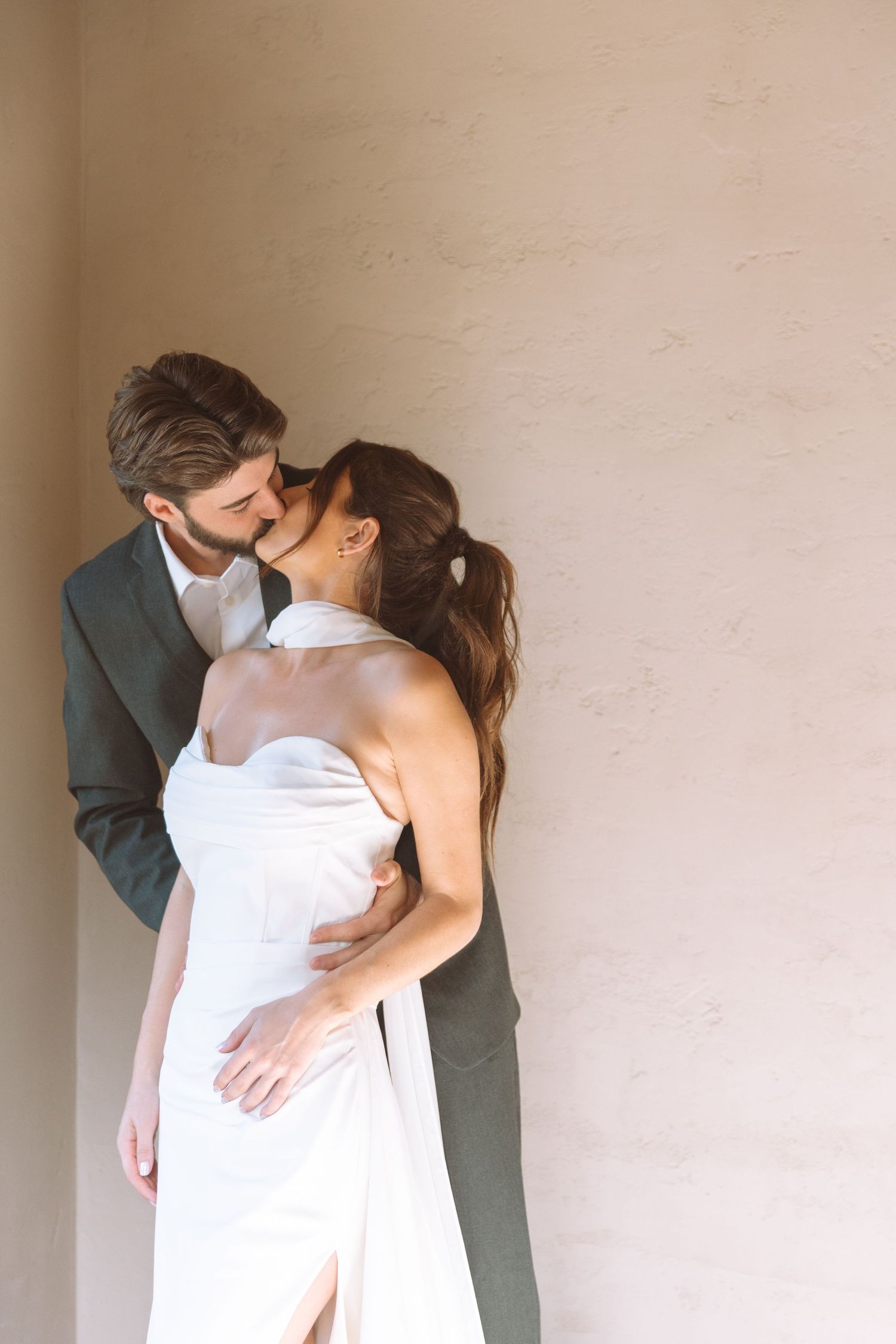 Bride and groom sharing a romantic kiss against a warm textured neutral wall.