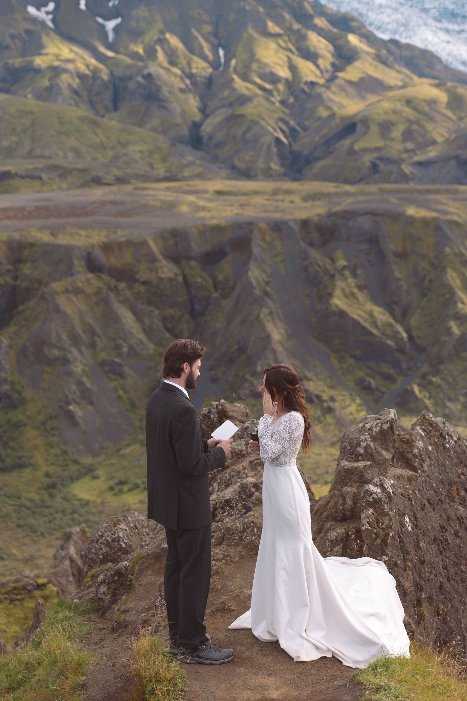 Bride and groom share intimate vow exchange on rugged Iceland cliffside with glacier mountain views.