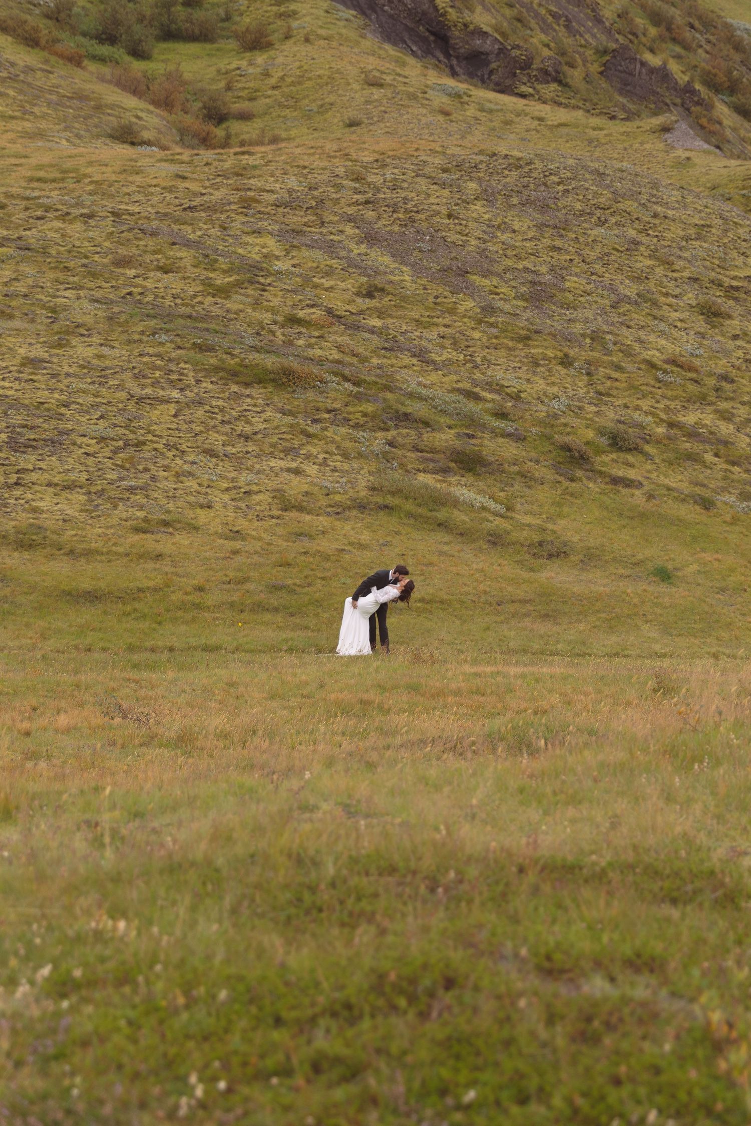 Bride and groom share a romantic dip kiss in vast open green hillside landscape during wedding shoot.
