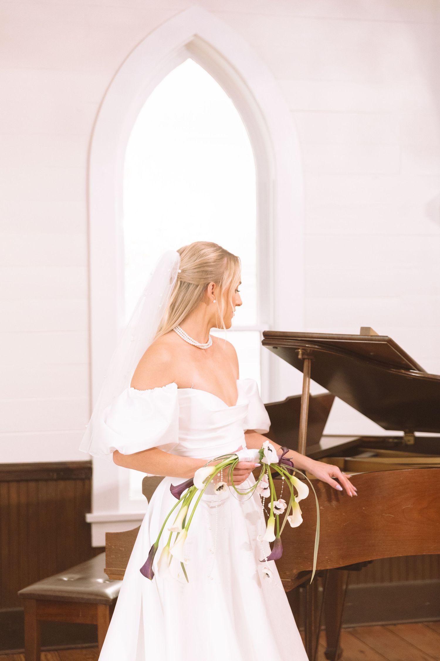 Blonde bride in off-shoulder wedding gown holding calla lily bouquet beside a grand piano.