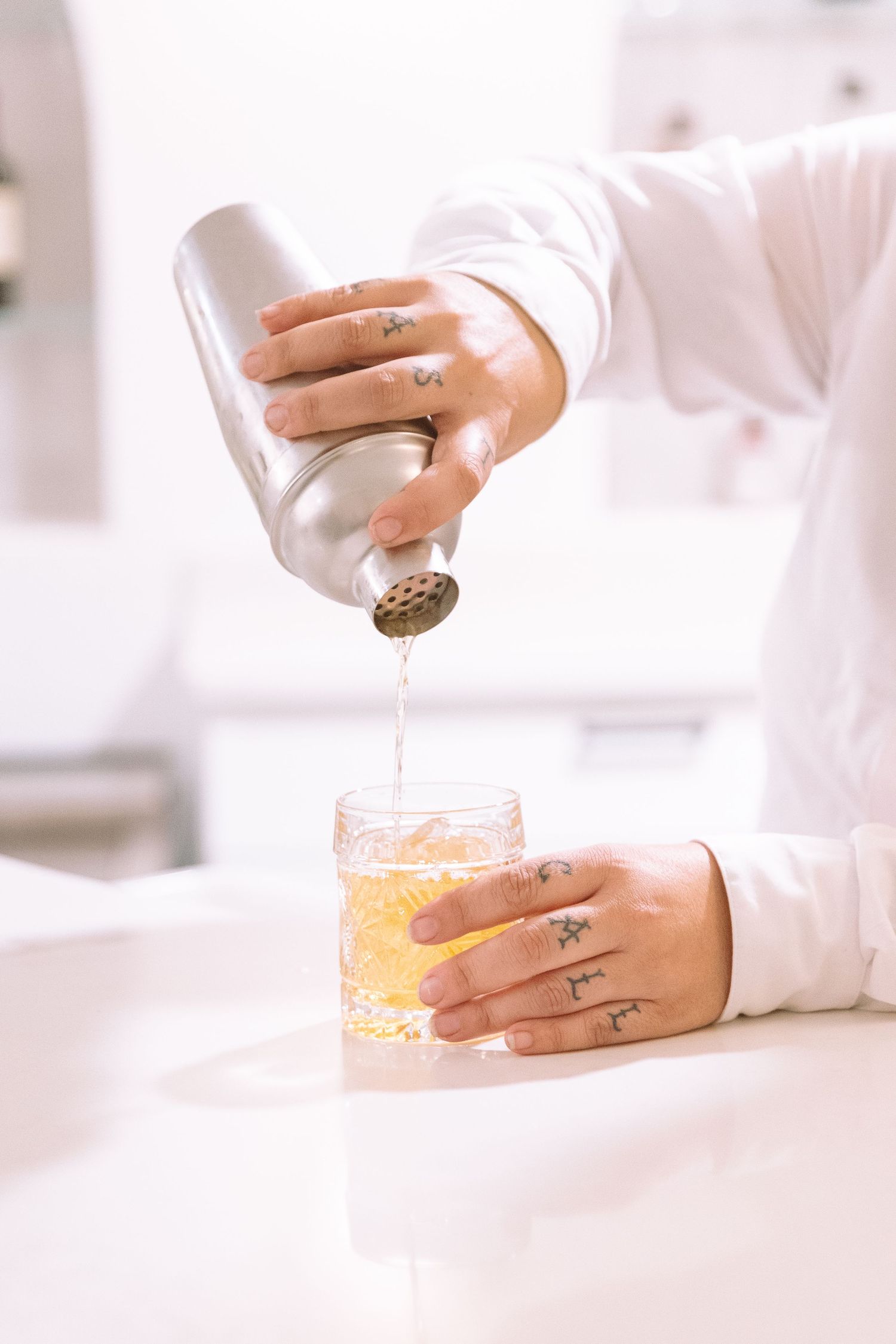 Tattooed bartender pouring cocktail shaker into an old fashioned glass on a white bar.