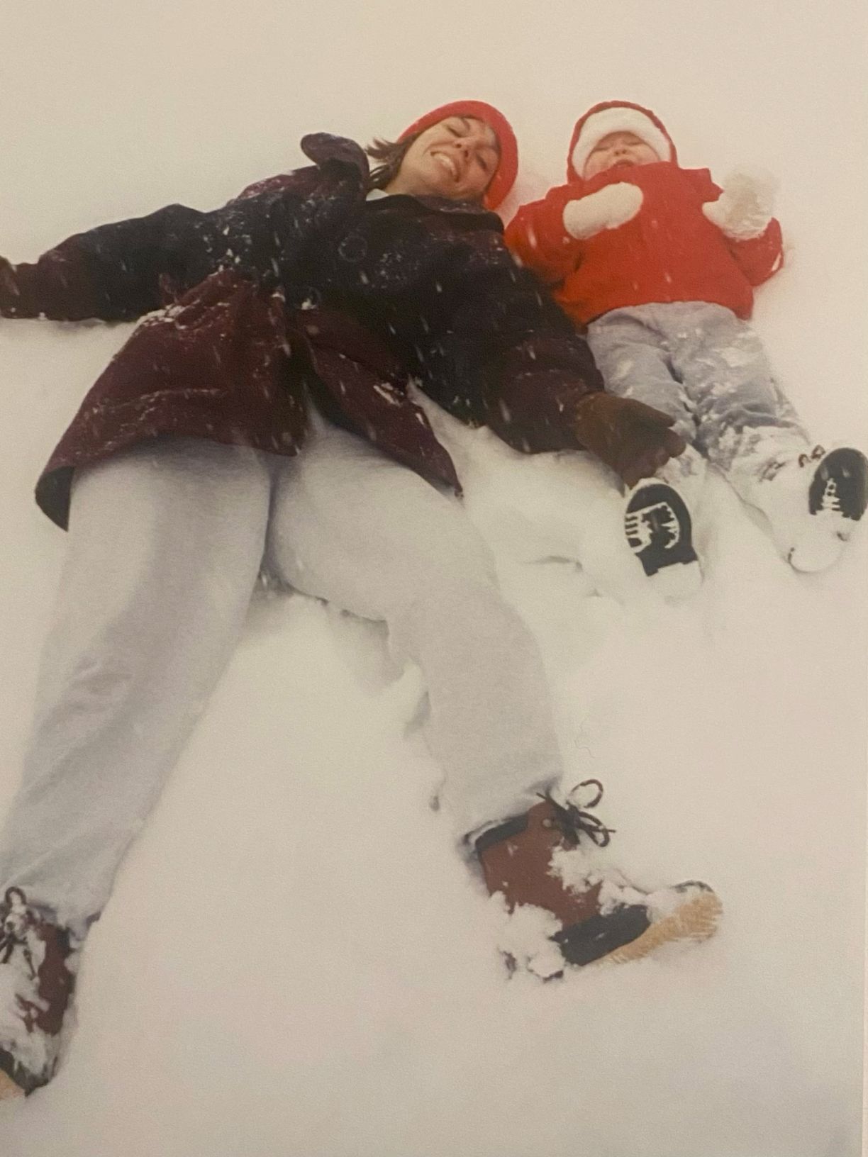 Mother and young child making snow angels together in fresh white snow during winter wearing colorful coats.