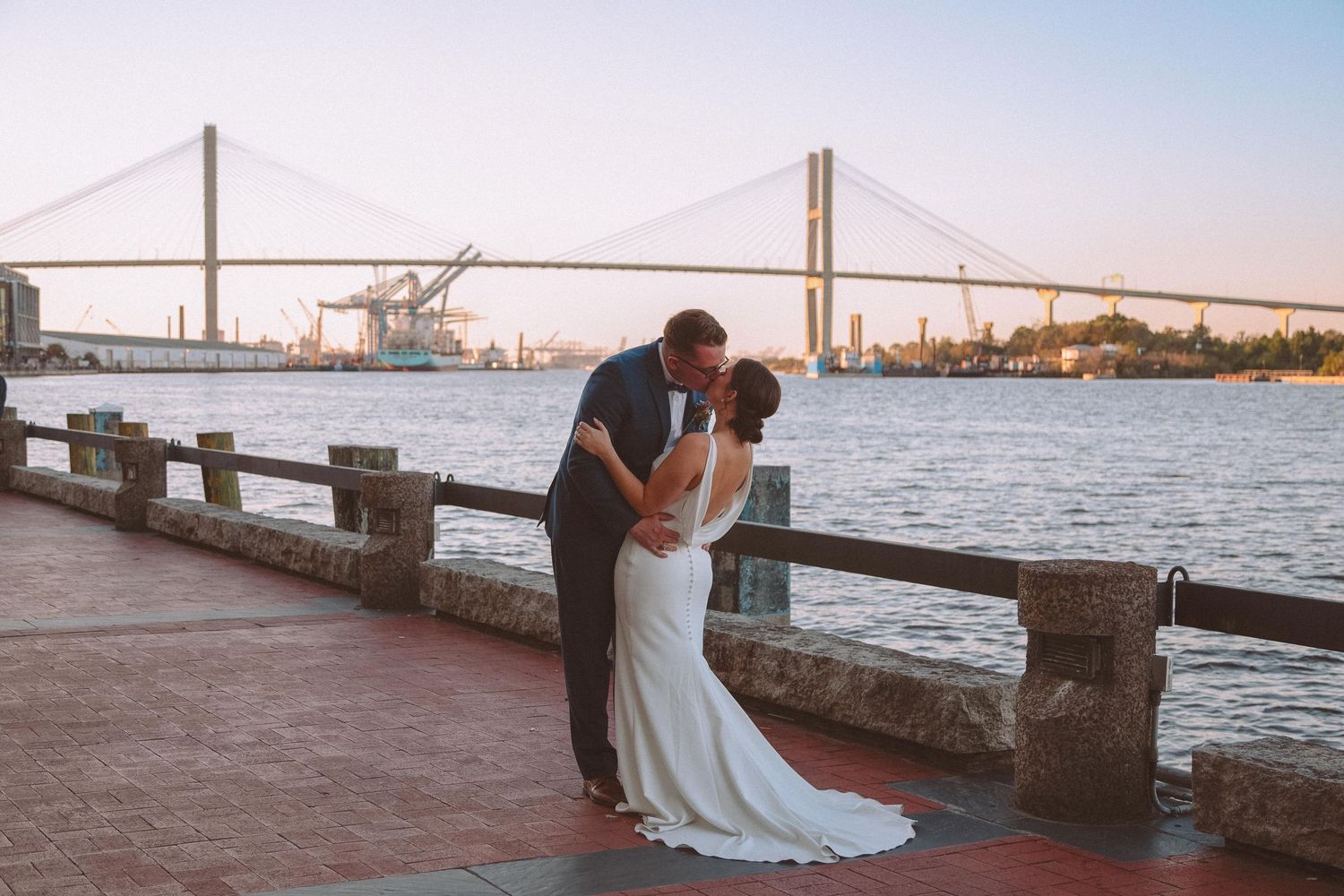 Newlyweds share romantic kiss on riverfront promenade with iconic cable-stayed bridge at sunset.