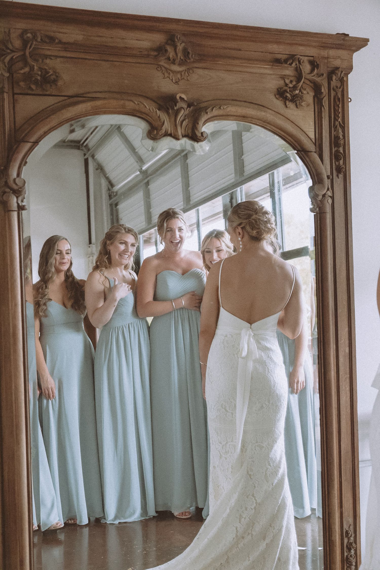 Bridesmaids in sage green gowns admire bride in lace dress reflected in ornate gold mirror.