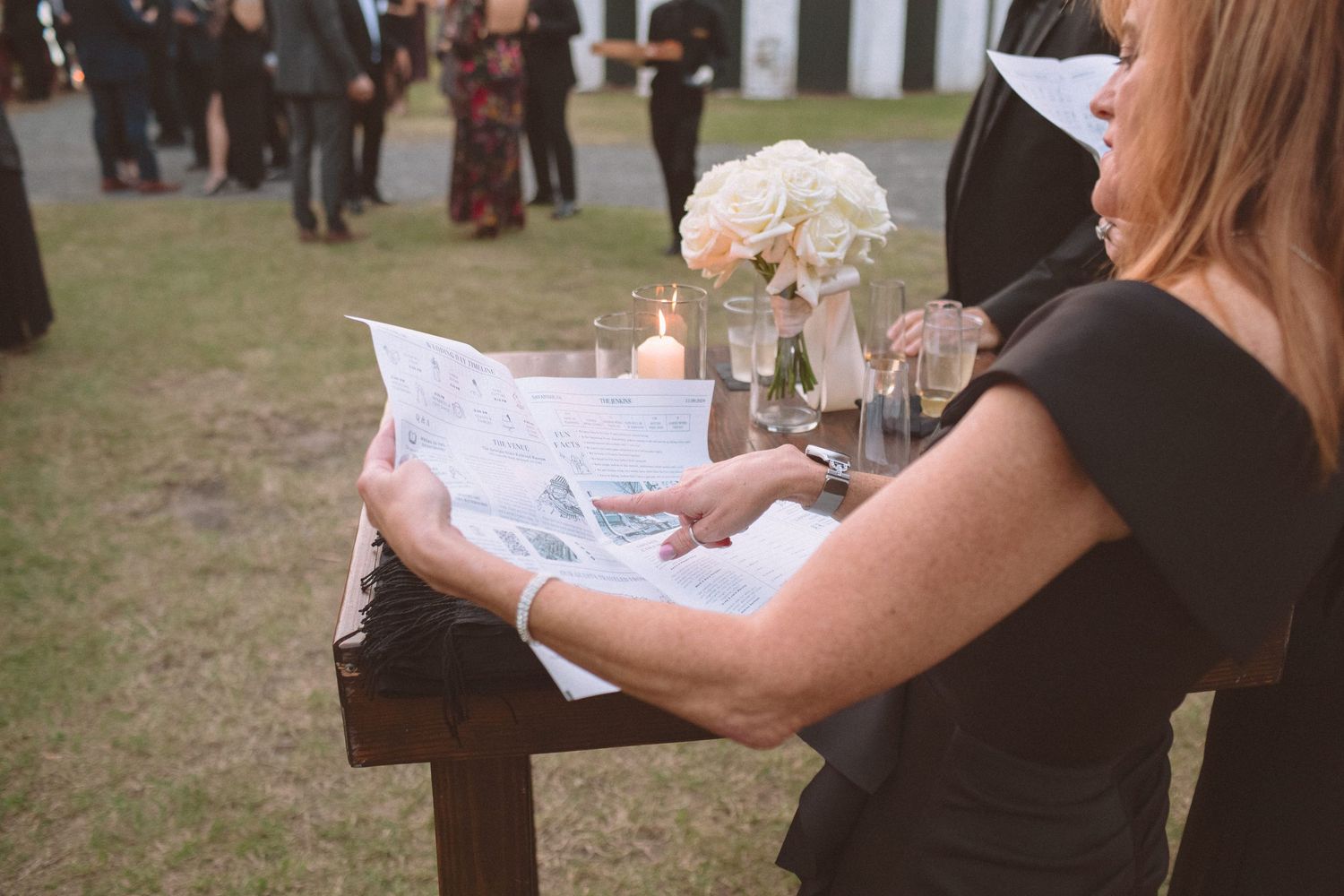 Wedding guest in black dress reading a newspaper-style program at an outdoor ceremony with floral decor.