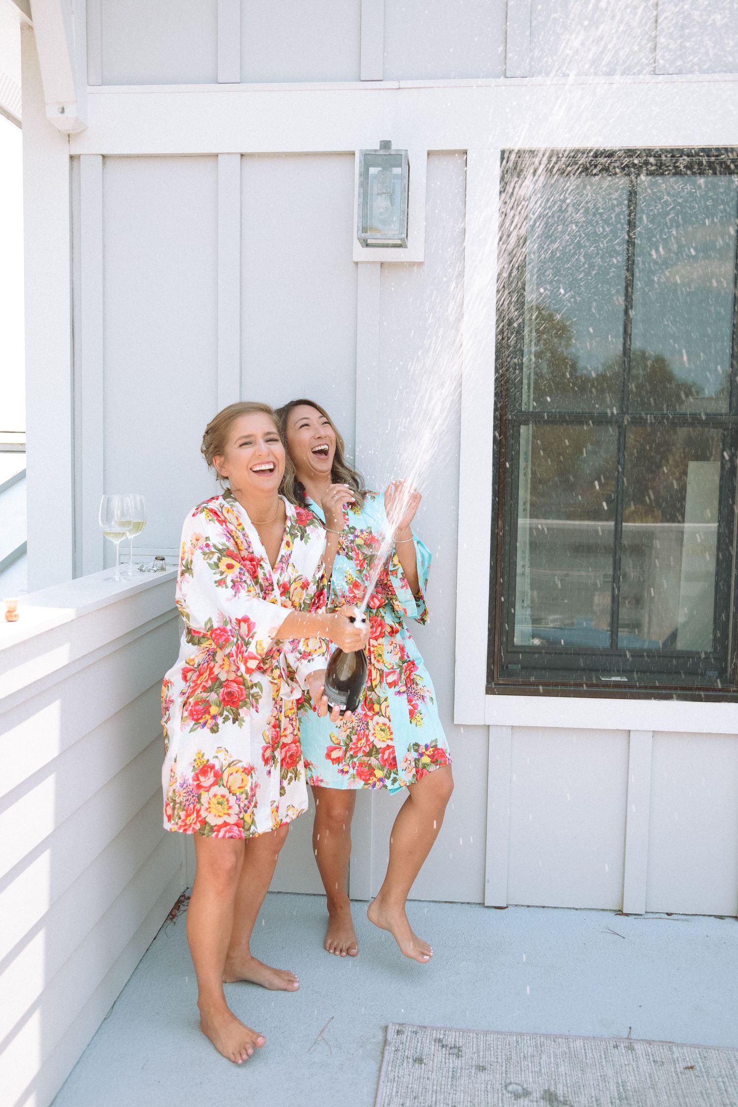 Two bridesmaids in colorful floral robes celebrating with champagne spray on a white deck.
