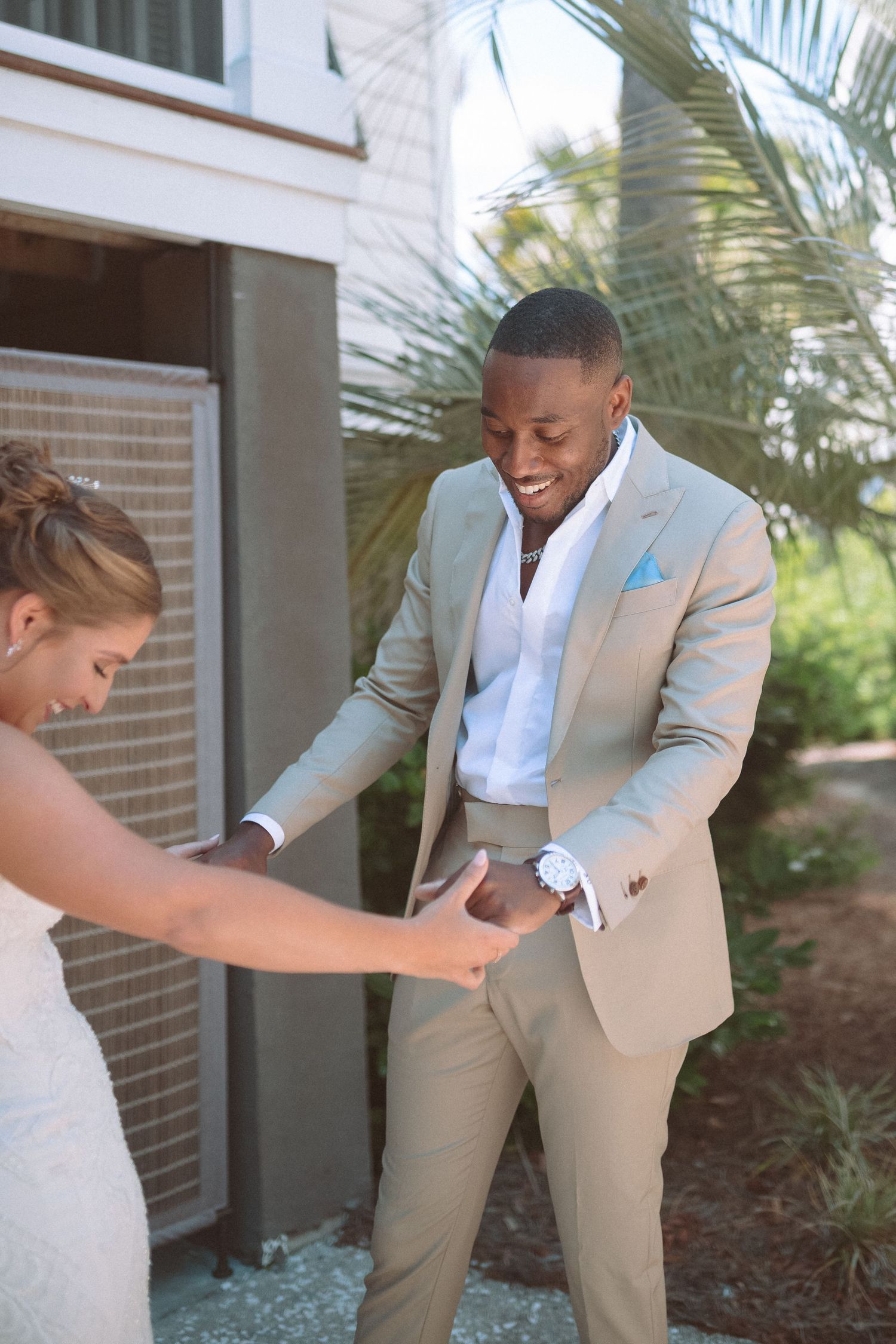 Smiling groom in beige suit holds bride's hands during first look wedding moment beside palm trees.